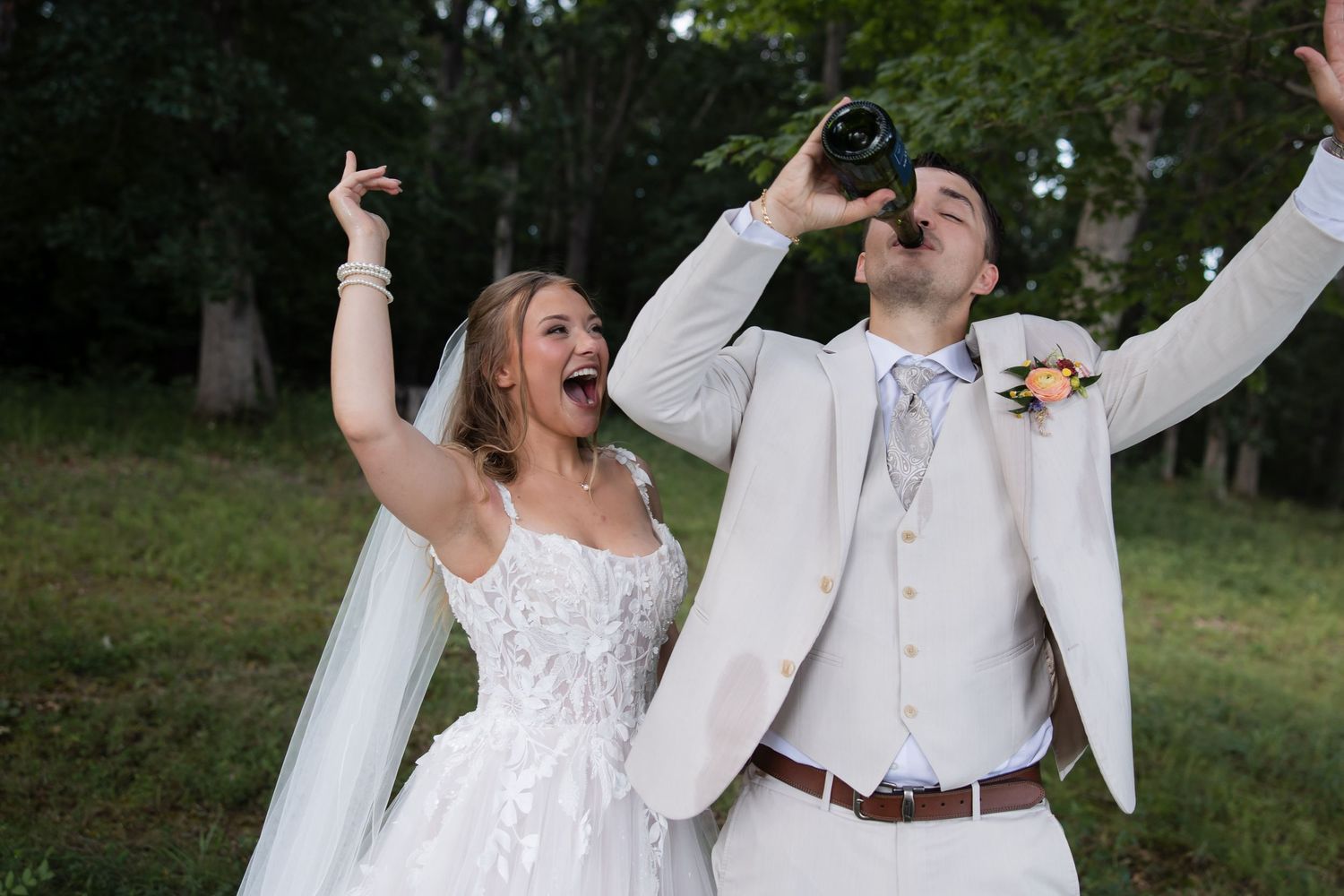 Joyful bride and groom celebrating on wedding day, groom drinking from bottle, bride cheering with arms raised outdoors.