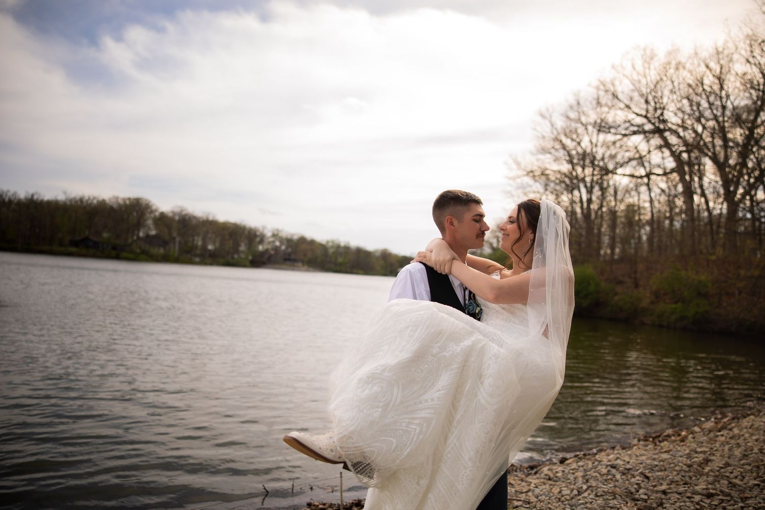 Groom carries bride in white gown and veil by a lake shore on their wedding day.