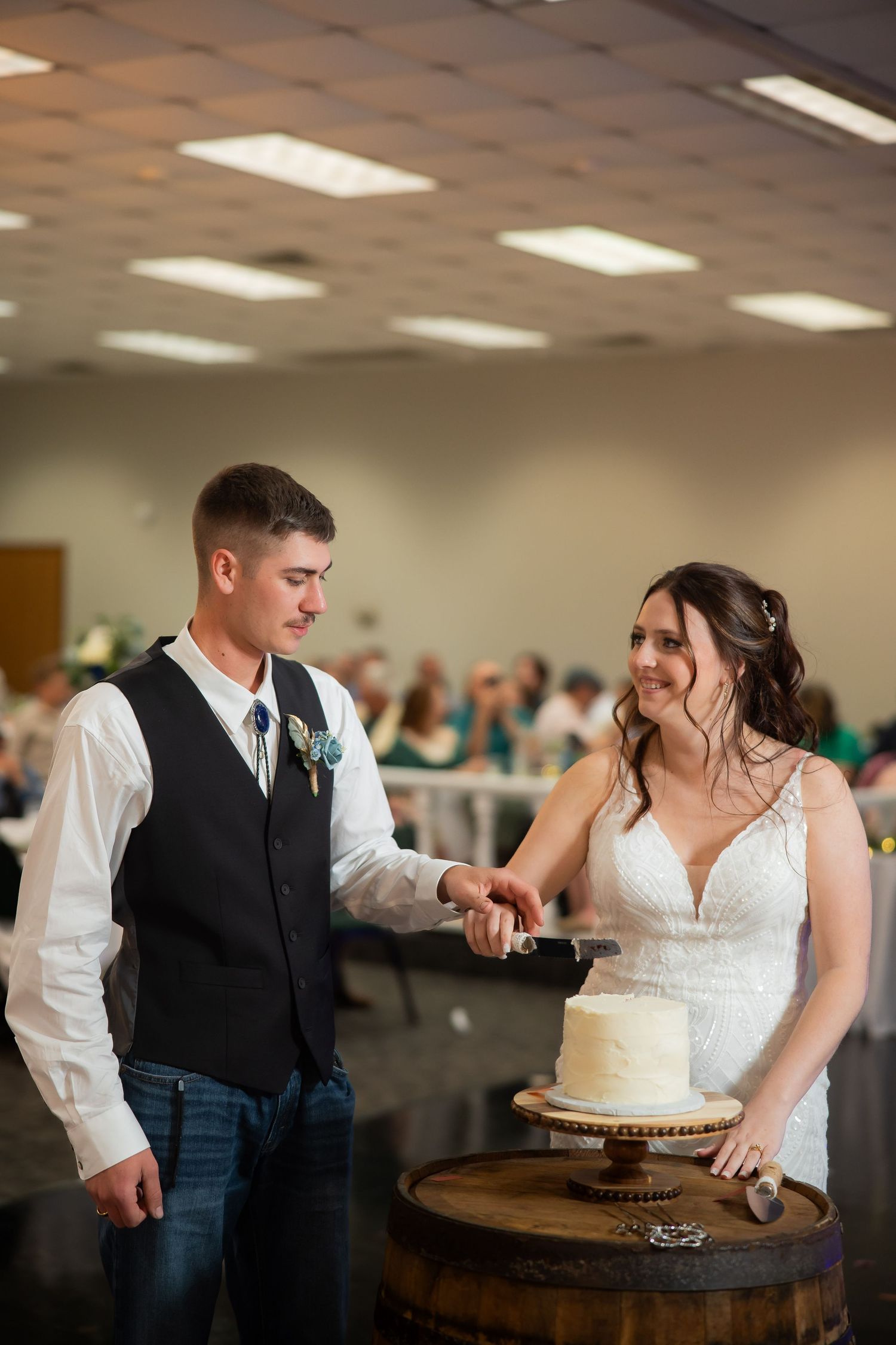 Bride and groom cut white wedding cake on rustic barrel at reception.
