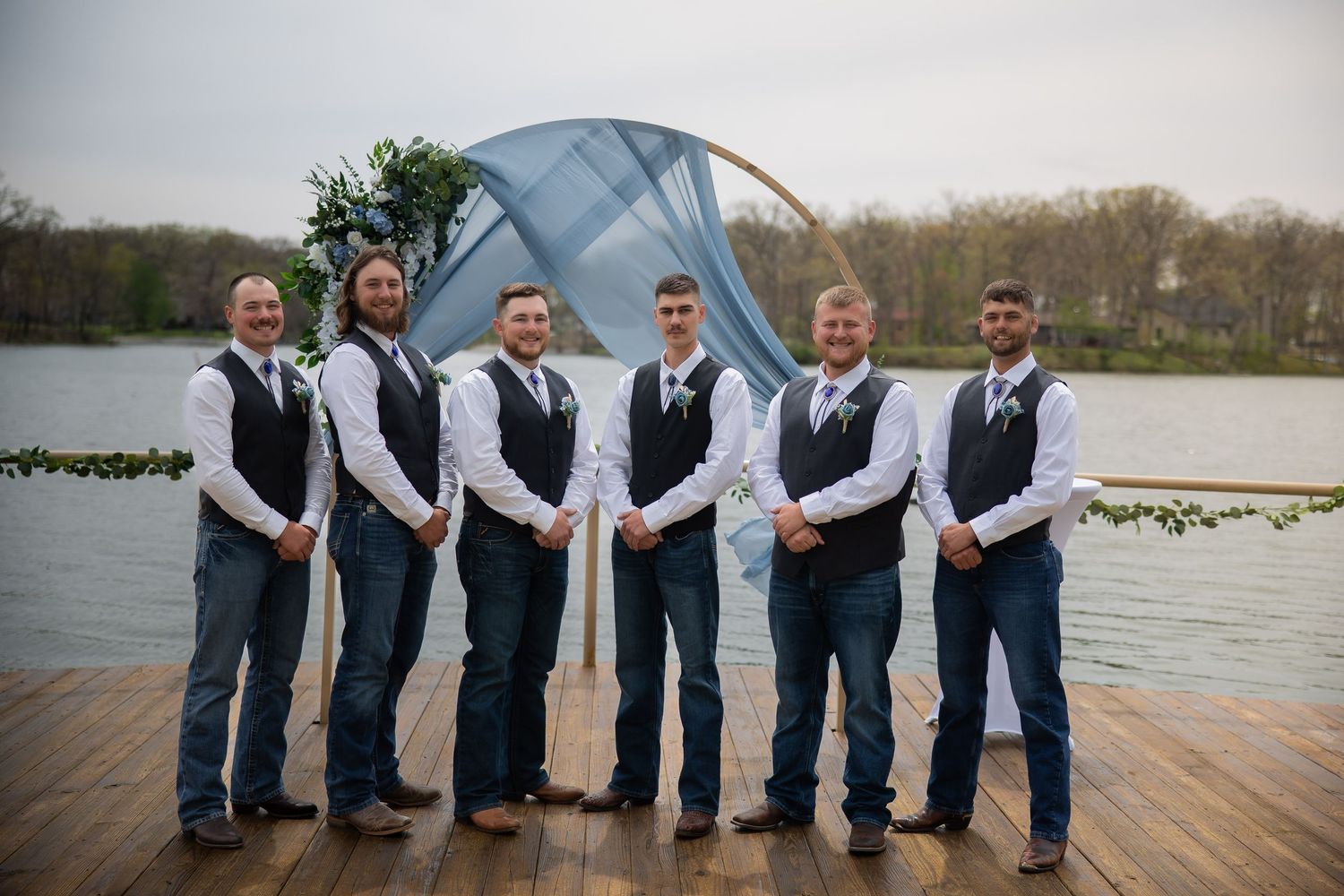 Six groomsmen in matching black vests and ties pose on a wooden dock by a lake at an outdoor wedding.