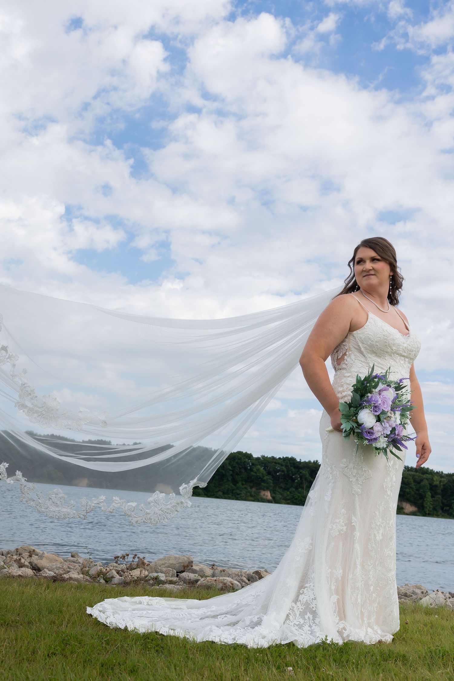 Bride in lace gown poses lakeside, veil billowing in wind, holding purple and white bouquet.