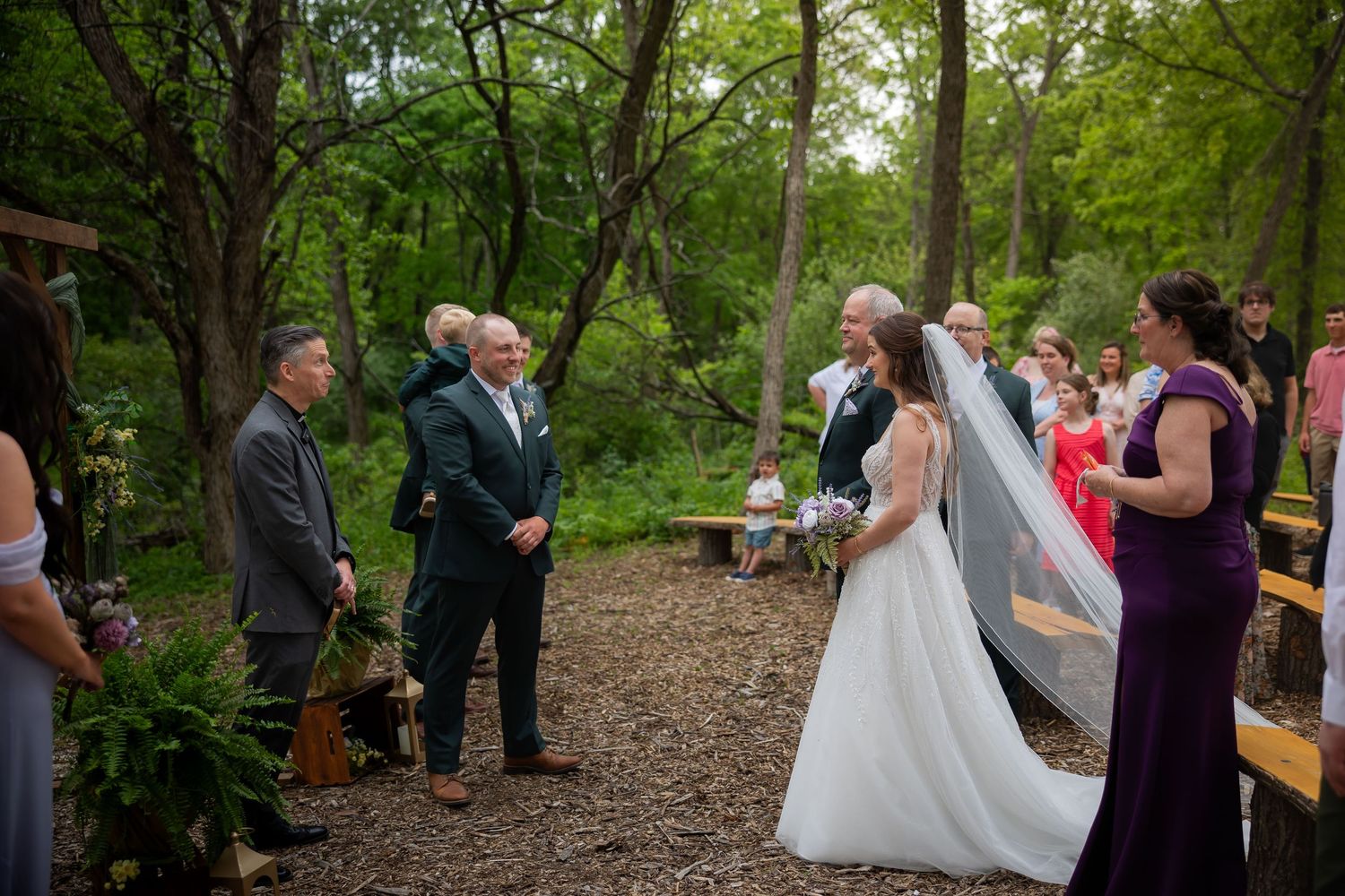 Bride in white gown and veil arrives at altar in lush green outdoor forest wedding ceremony.