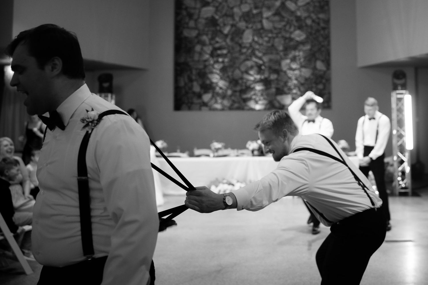 Black and white photo of boys in suspenders playfully pulling each other at a formal wedding reception.