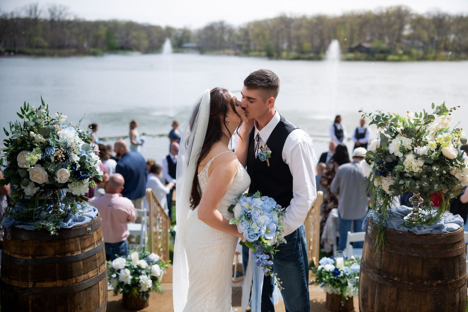 Bride and groom share first kiss at outdoor lakeside wedding ceremony with floral arrangements on barrels.