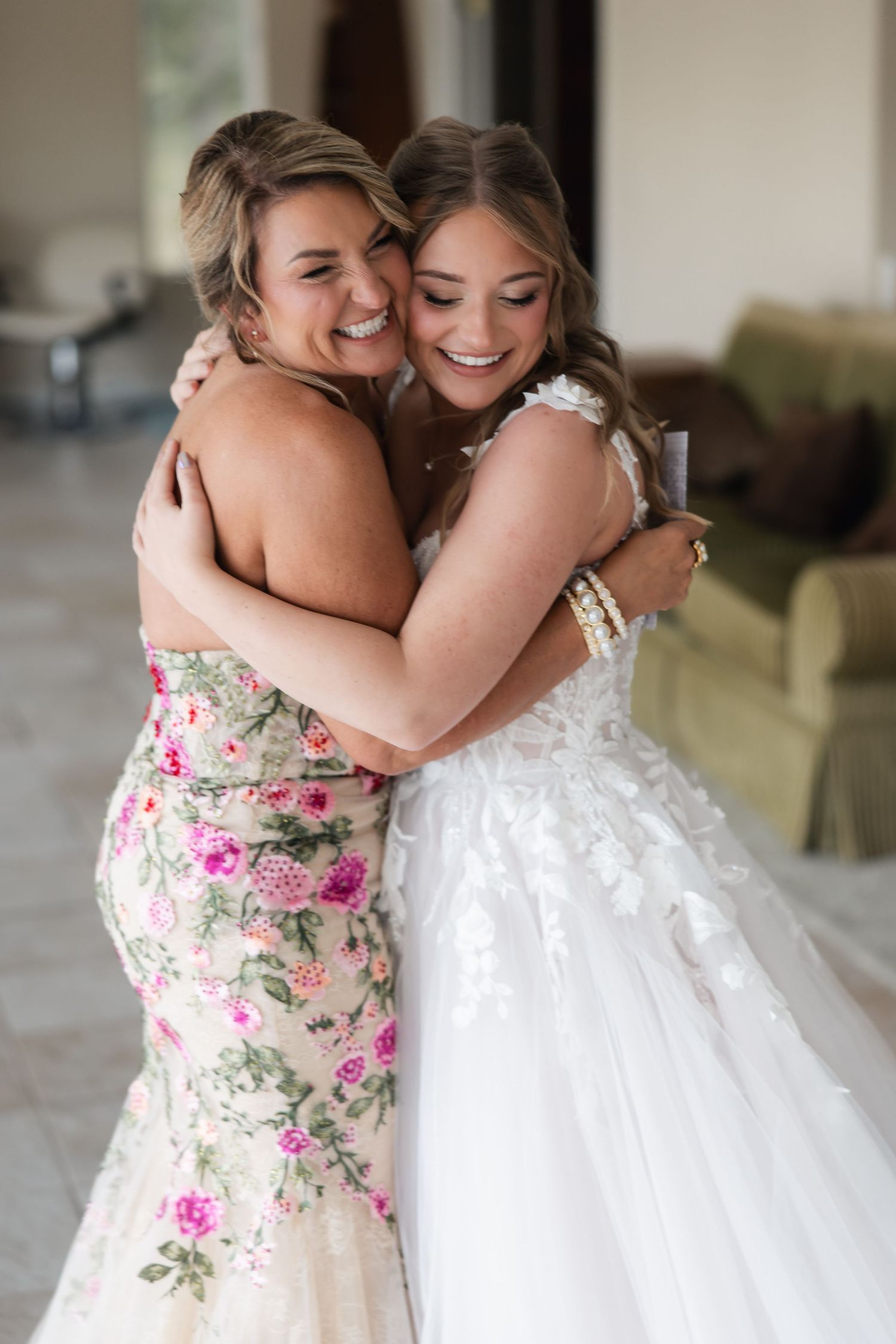 Bride in lace gown embraces woman in floral dress, both smiling joyfully.