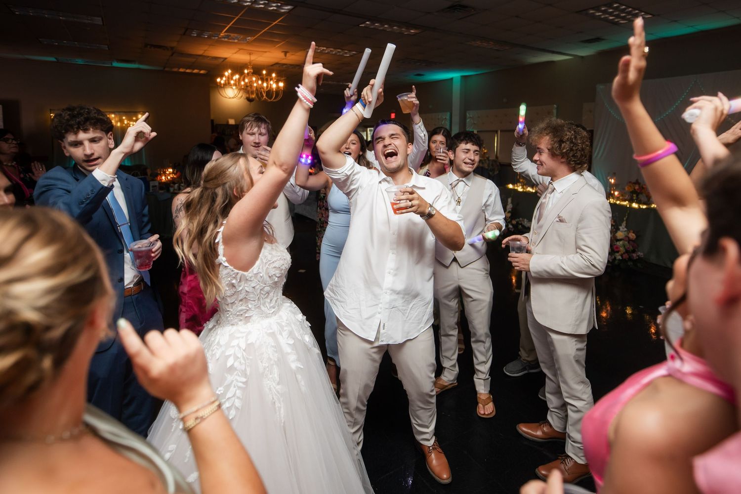 Bride and groom celebrate with guests on the dance floor at a lively indoor wedding reception.