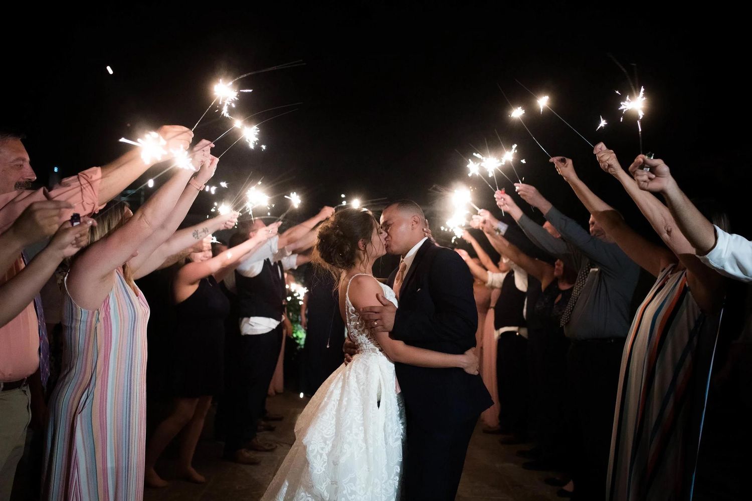 Bride and groom kissing under sparklers held by wedding guests during romantic nighttime sendoff ceremony.