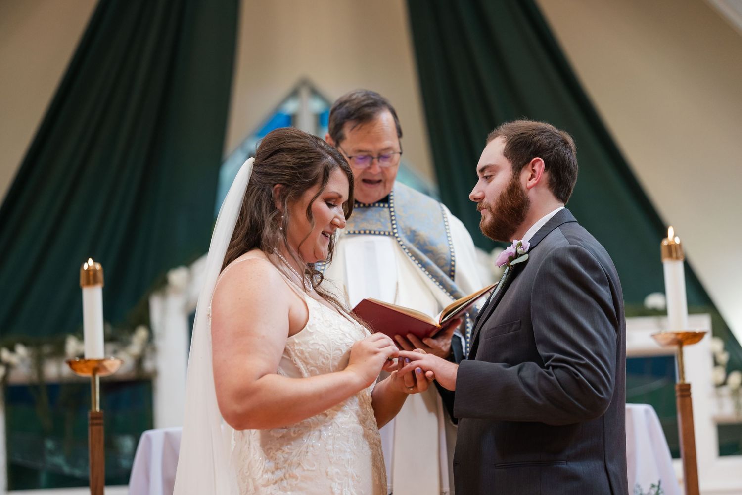 Bride and groom exchanging rings at altar with smiling officiant during church wedding ceremony.