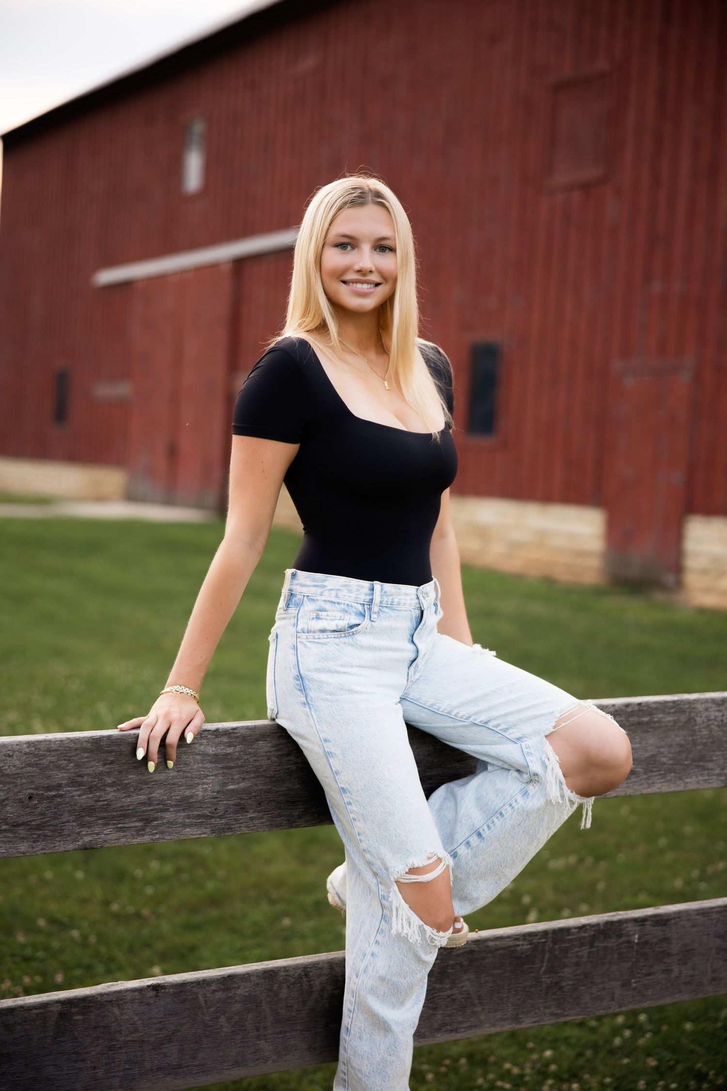 Figure in black top and ripped jeans posing casually on a wooden fence near a red barn.