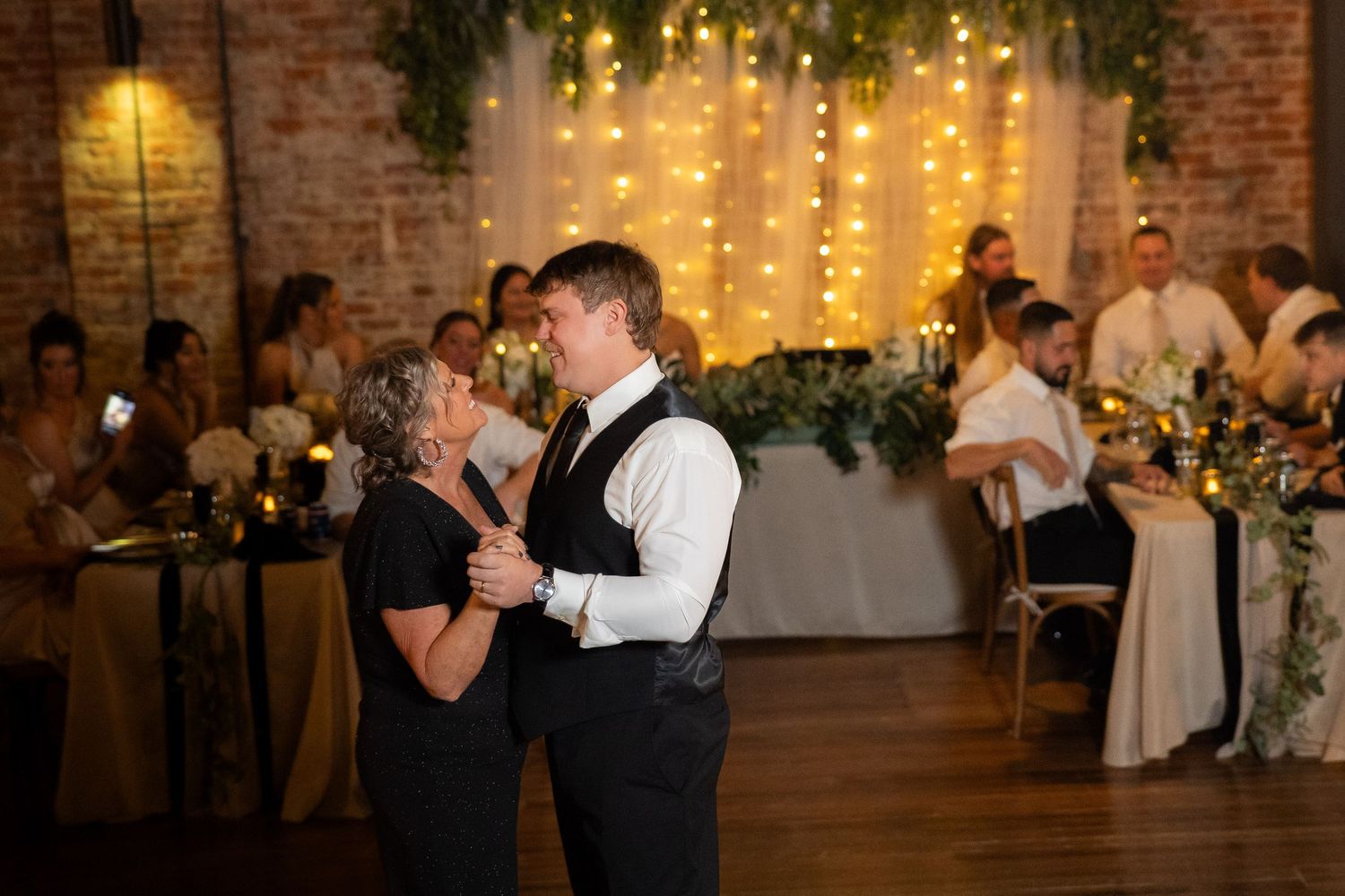 Wedding reception in rustic venue with string lights and greenery decorations.
