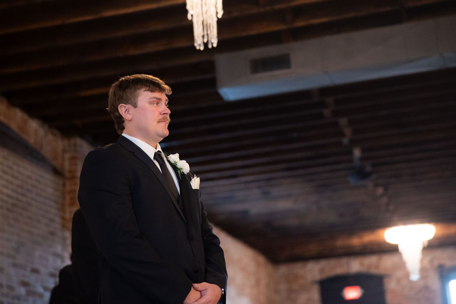 A groom in a black tuxedo stands at the altar during an indoor wedding ceremony.
