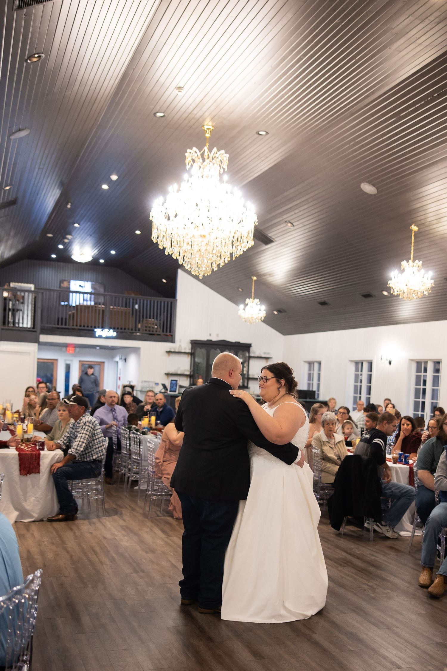 Wedding reception guests gather in an elegant ballroom with crystal chandeliers and white decor for a celebration.