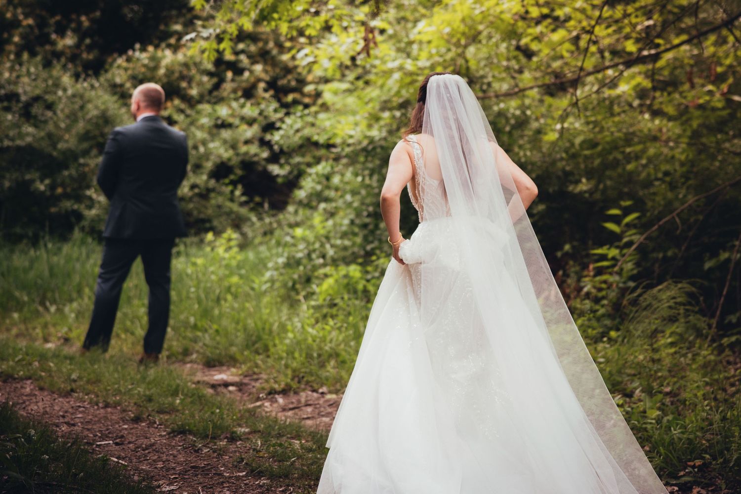 Bride in white gown and veil walks toward groom in navy suit on a wooded garden path.
