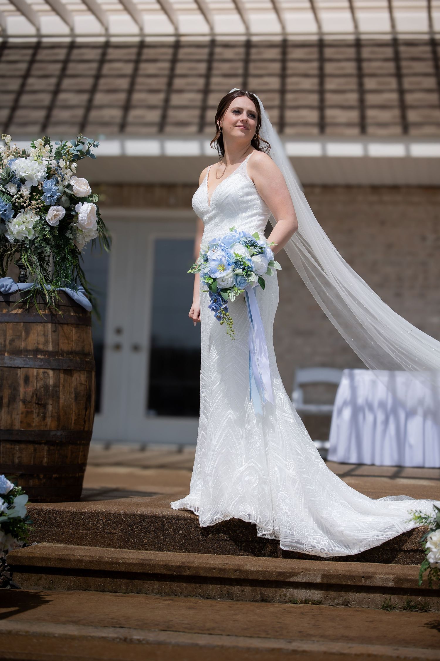 Elegant bride in a white lace gown holding a blue and white bouquet with a flowing veil outdoors.