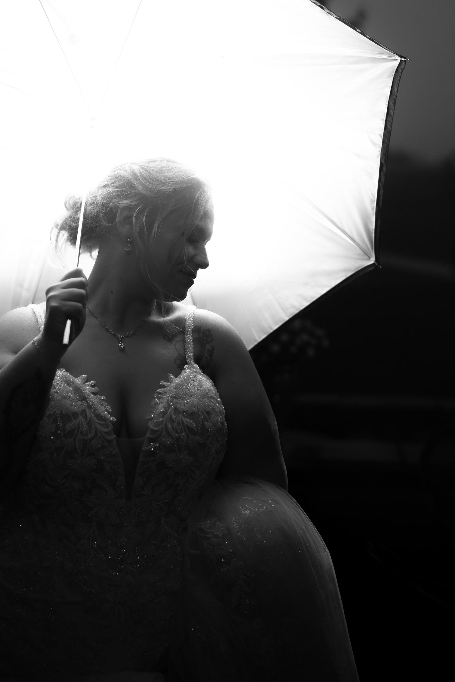 Black and white photo of a bride in a lace gown holding a white umbrella, backlit with dramatic lighting.