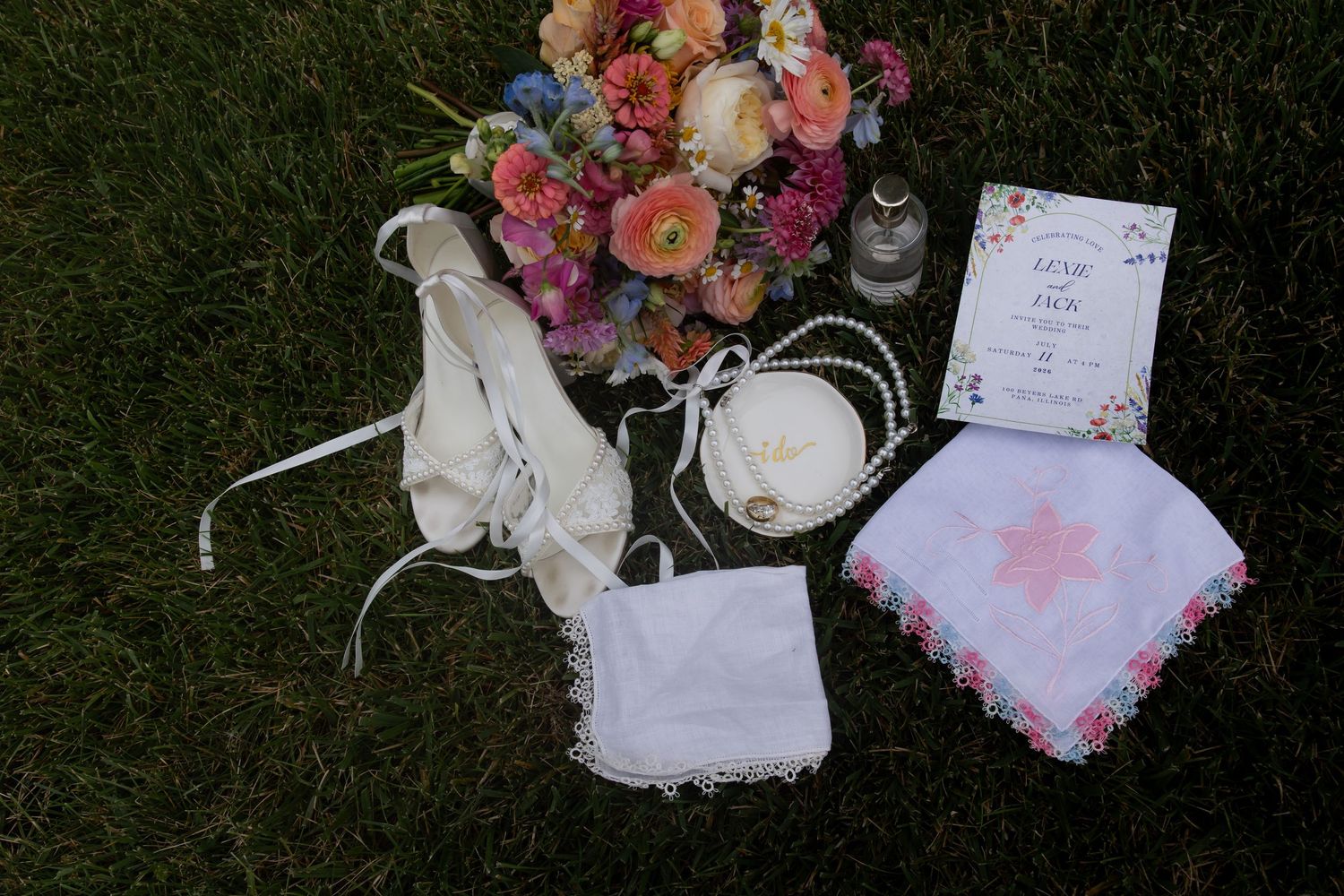 Wedding flat lay with bridal accessories, rings, handkerchief, invitation, pearls, and colorful floral bouquet on green grass.