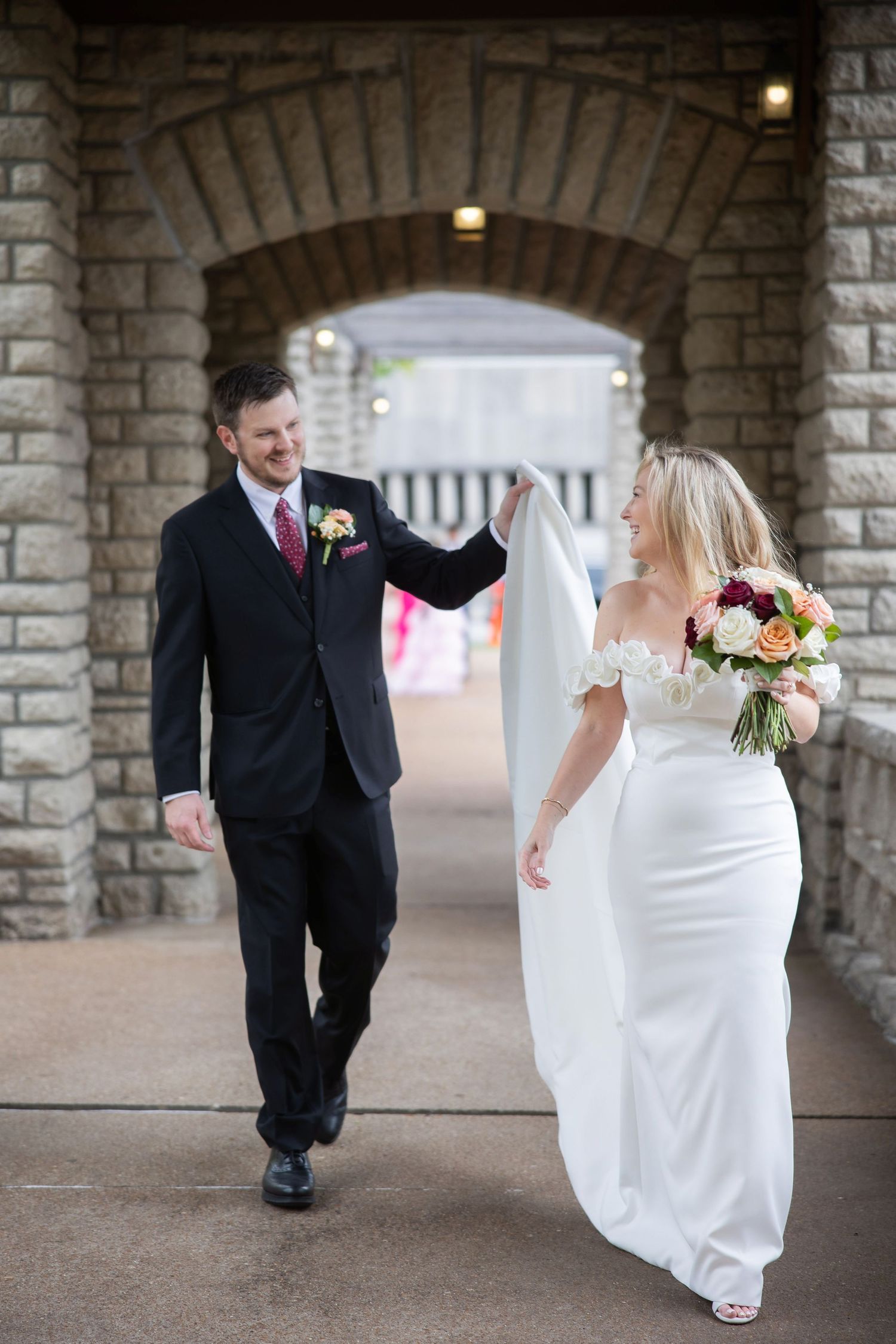 Couple walking and playing under stone archway during wedding photoshoot.