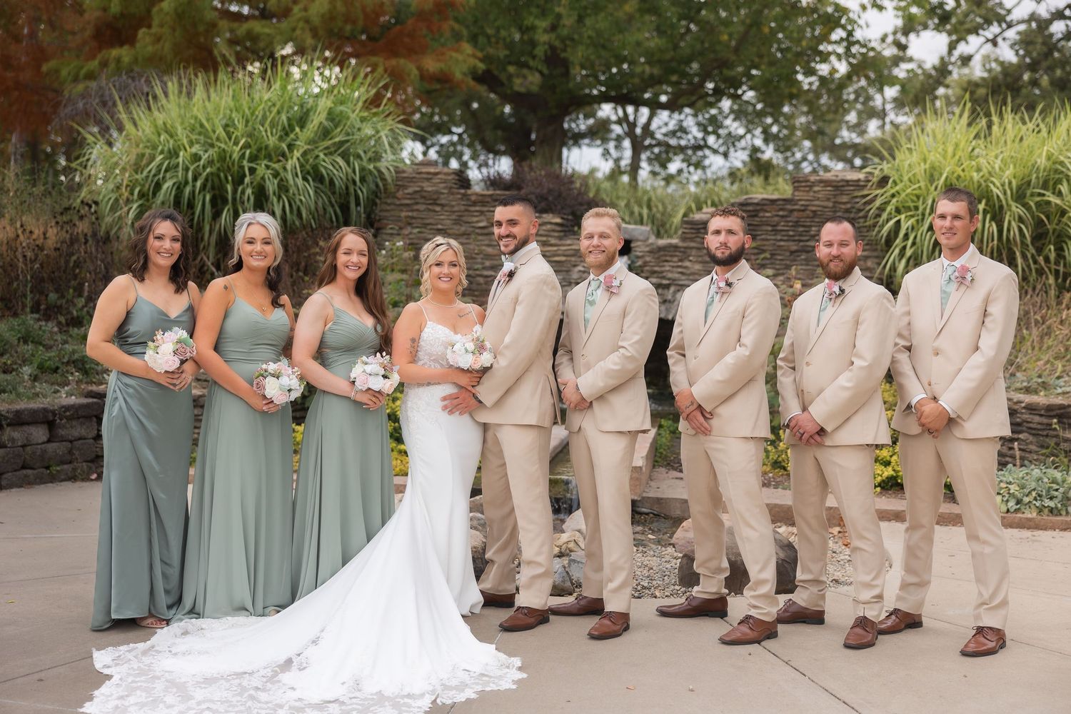 Wedding party poses outdoors with bridesmaids in sage green dresses and groomsmen in tan suits.
