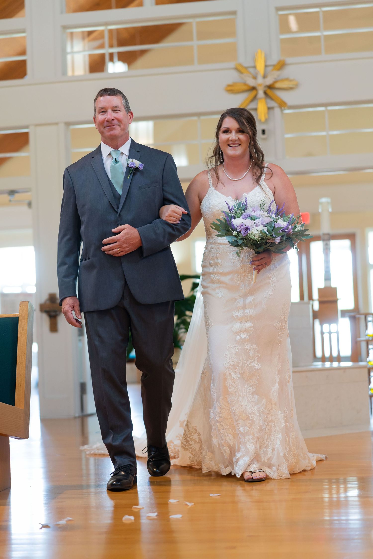 Bride in lace gown holding purple bouquet walks down church aisle with escort in gray suit at wedding ceremony.