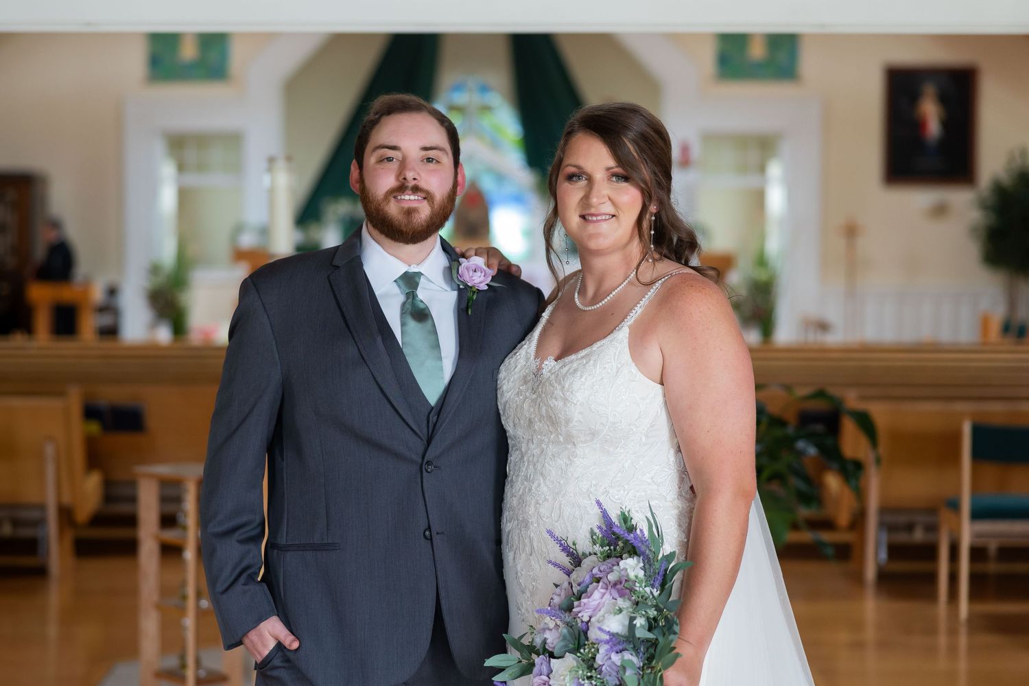 Bride and groom posing together in a church, bride holding purple bouquet in white lace gown, groom in gray suit.