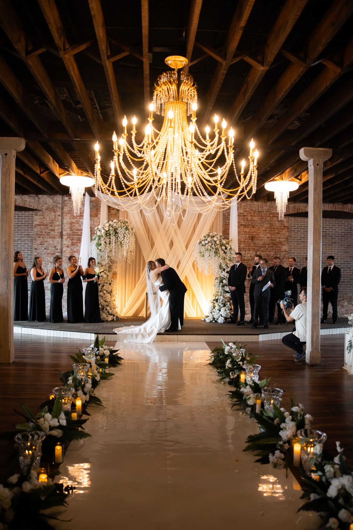 Elegant indoor wedding ceremony with chandelier, floral aisle, candles, and couple kissing at altar.