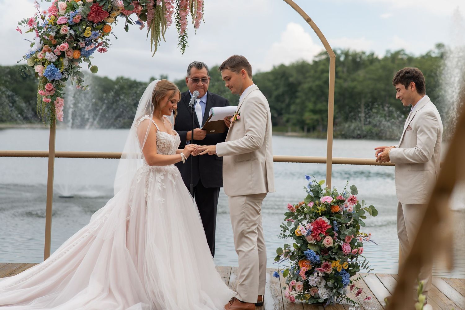 Couple exchanges rings at waterfront wedding ceremony under floral arch with fountain in background.