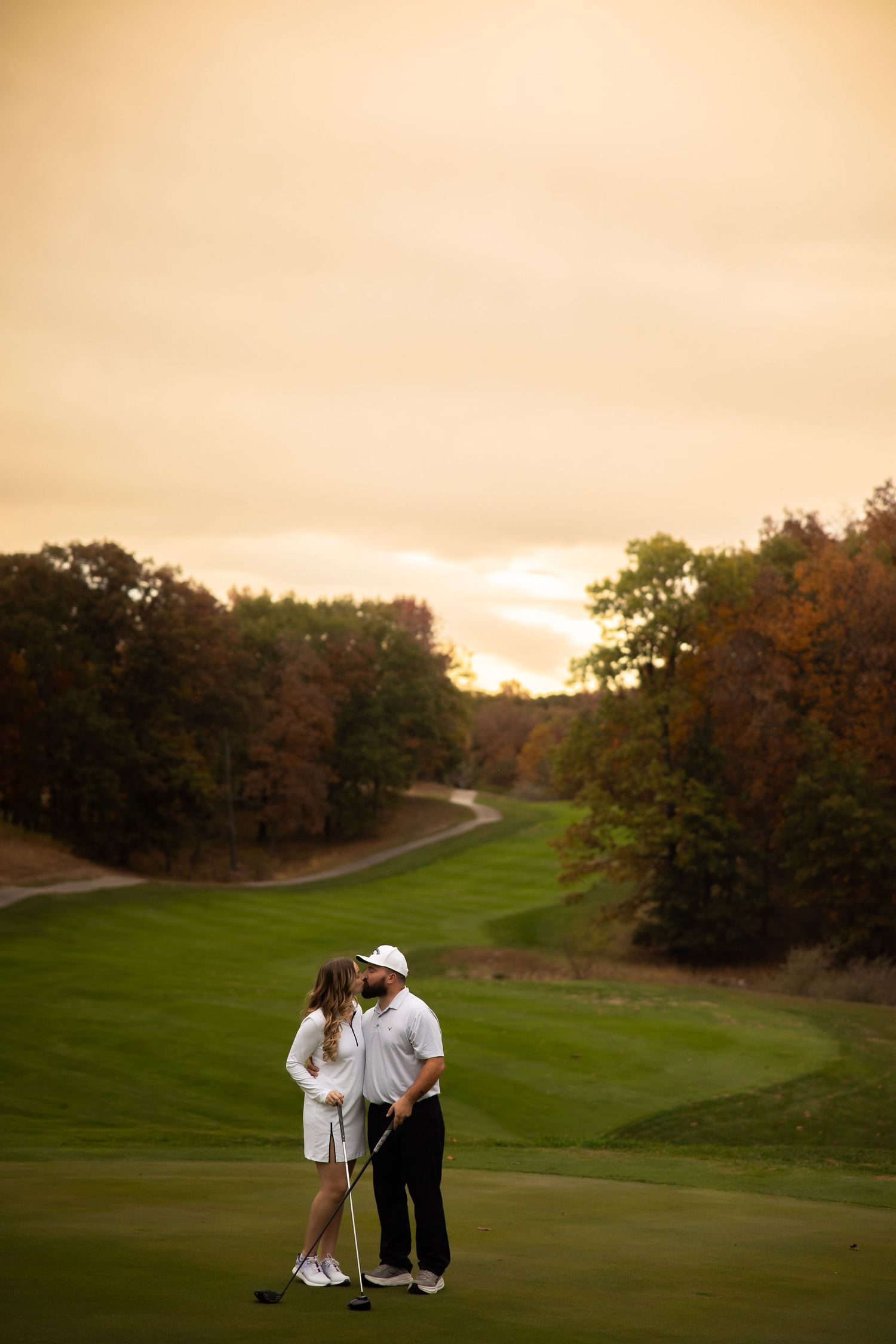 Two golfers embrace on a scenic golf course at sunset with autumn trees and winding fairway in the background.