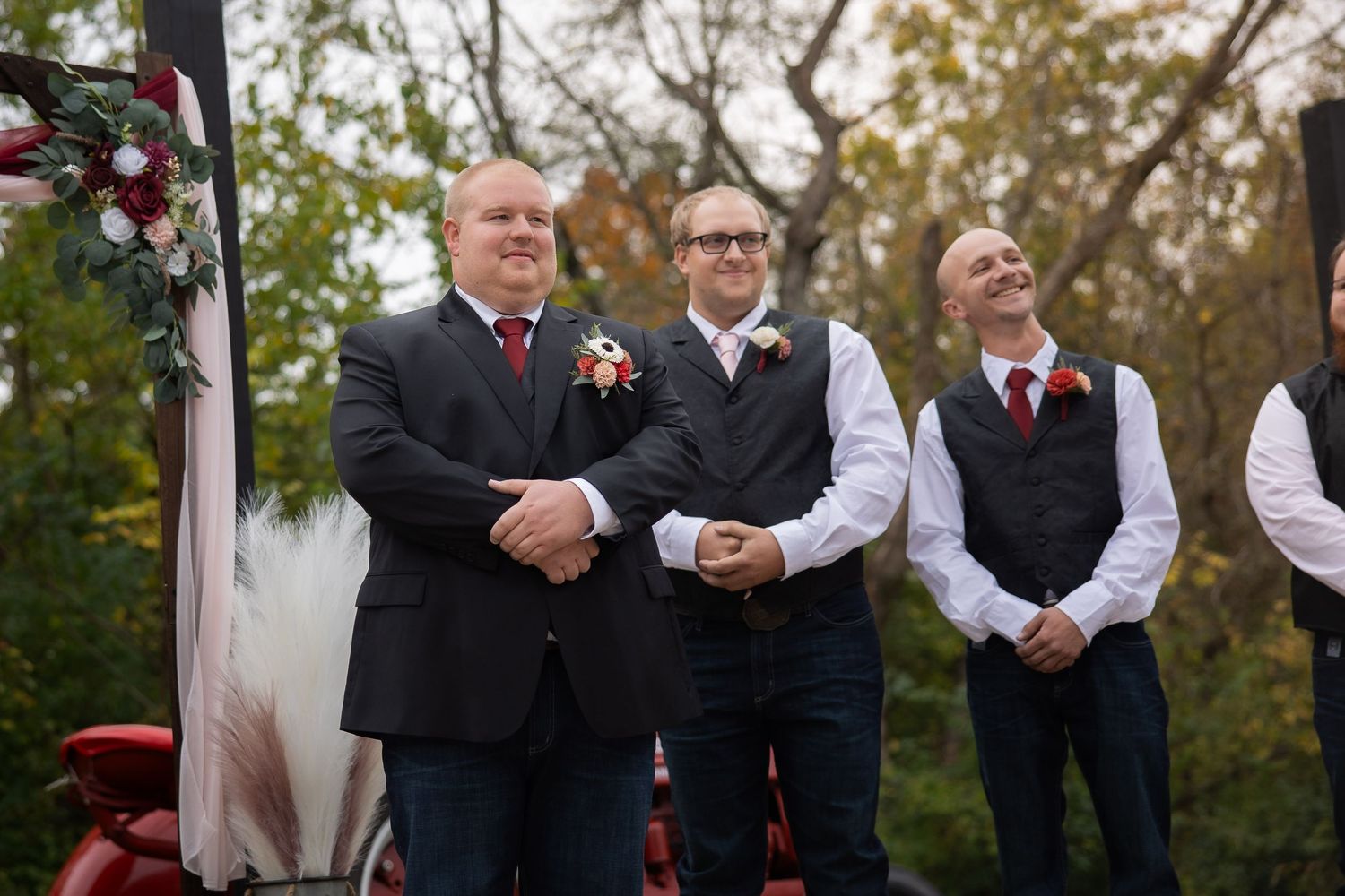 Groomsmen stand together during an outdoor autumn wedding ceremony near a decorated floral arch.