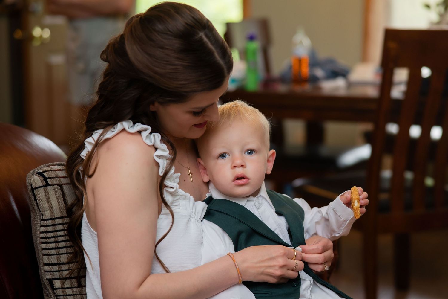 A young mother in a white dress holds a blue-eyed baby boy in a green vest at a wedding reception.