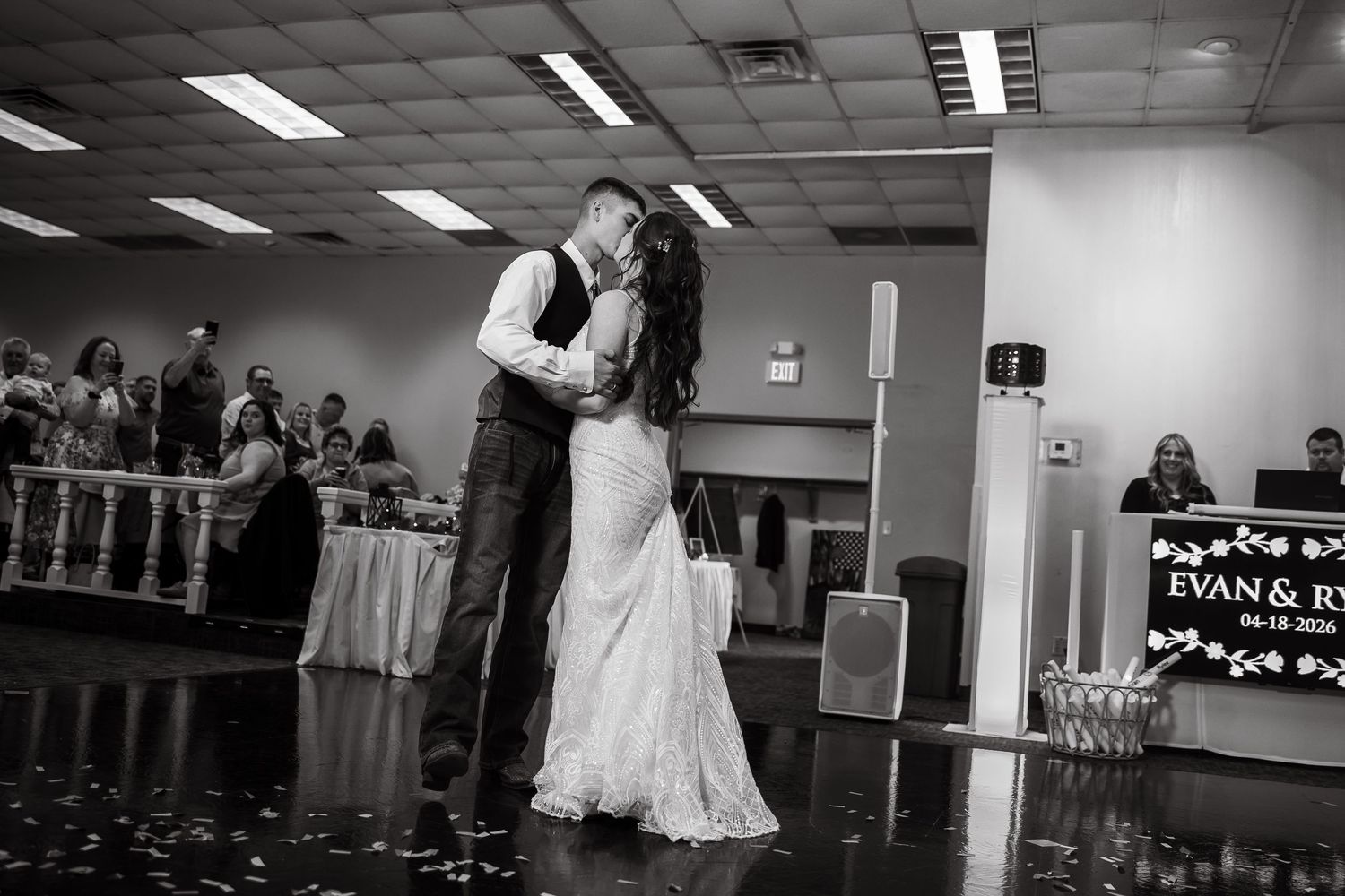Black and white photo of bride and groom sharing first dance at wedding reception with guests watching in background.