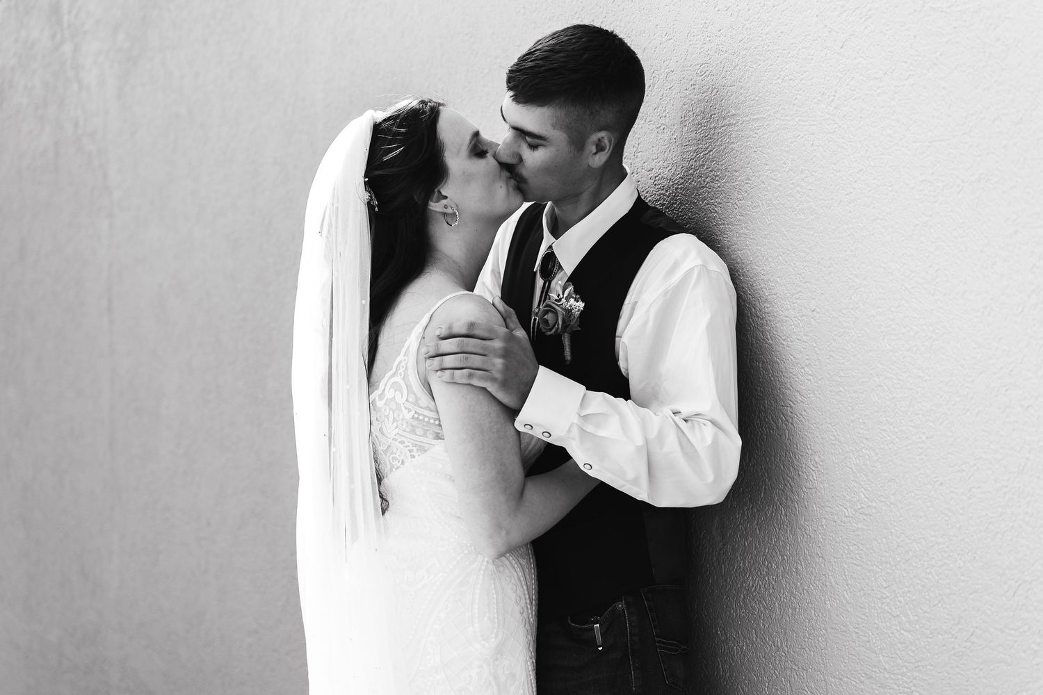 Black and white photo of a bride and groom sharing a romantic kiss against a wall on their wedding day.