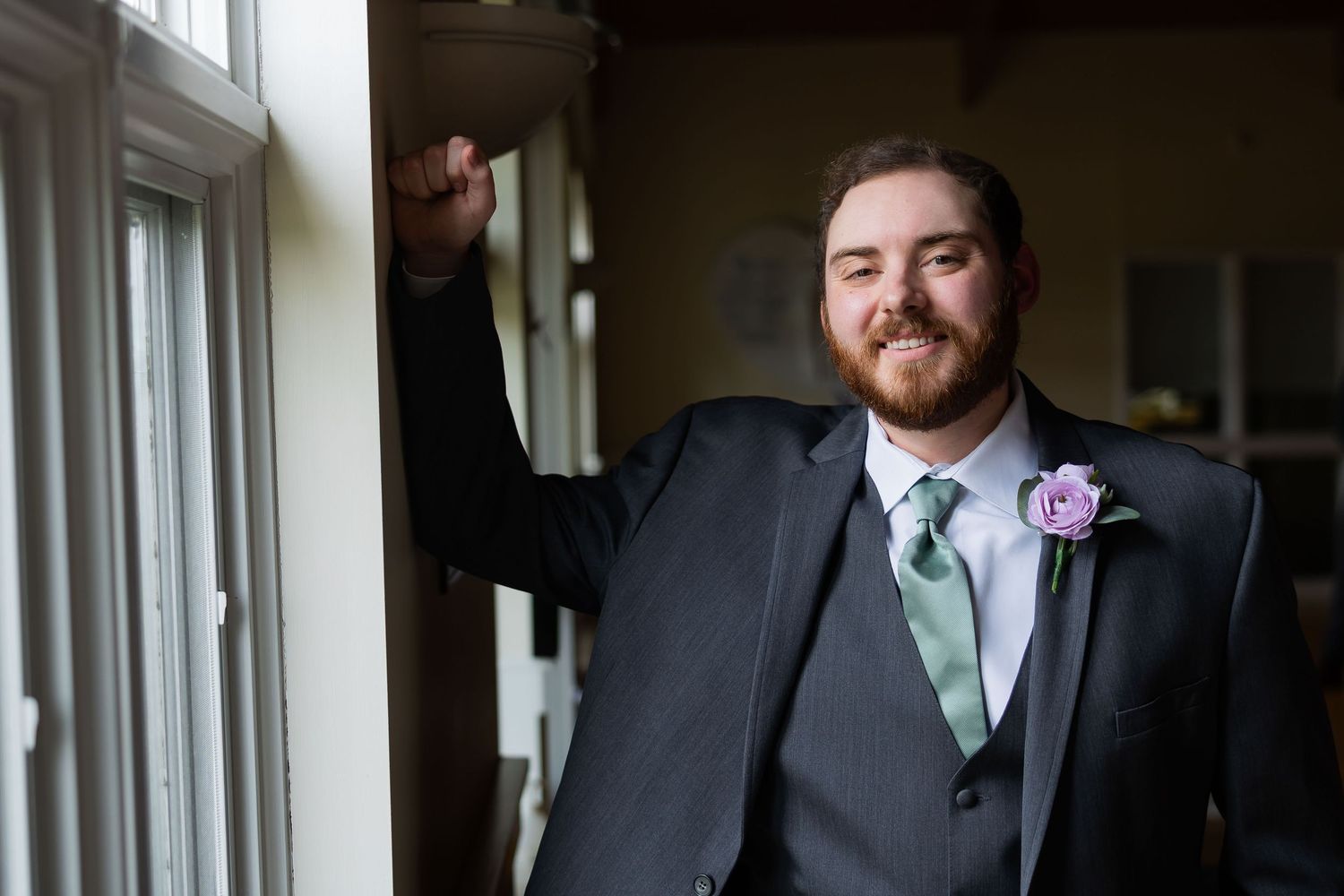 Groom in gray suit with mint tie and pink boutonniere smiles while leaning against a window indoors.