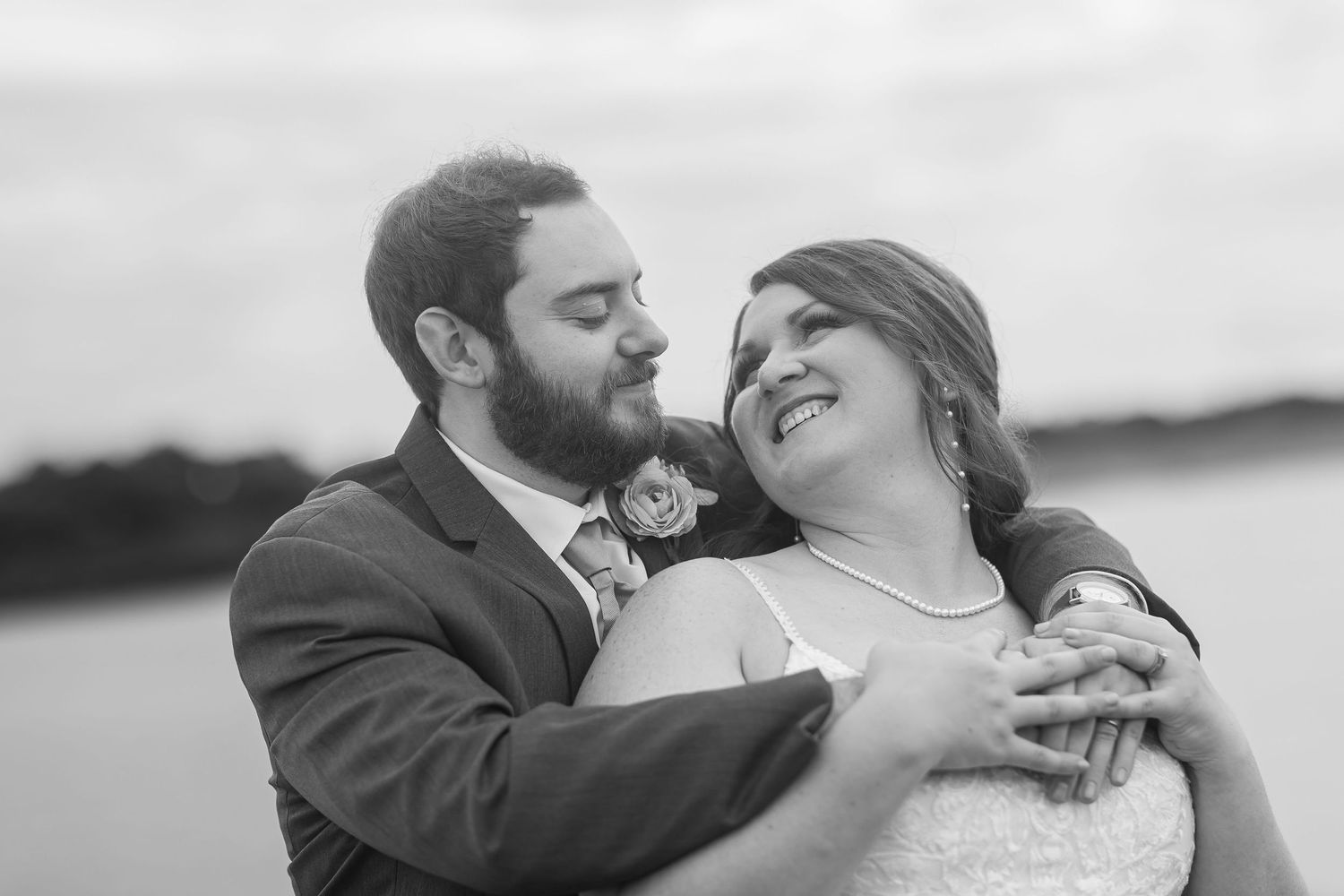 Black and white wedding photo of smiling bride and bearded groom embracing near a lake.