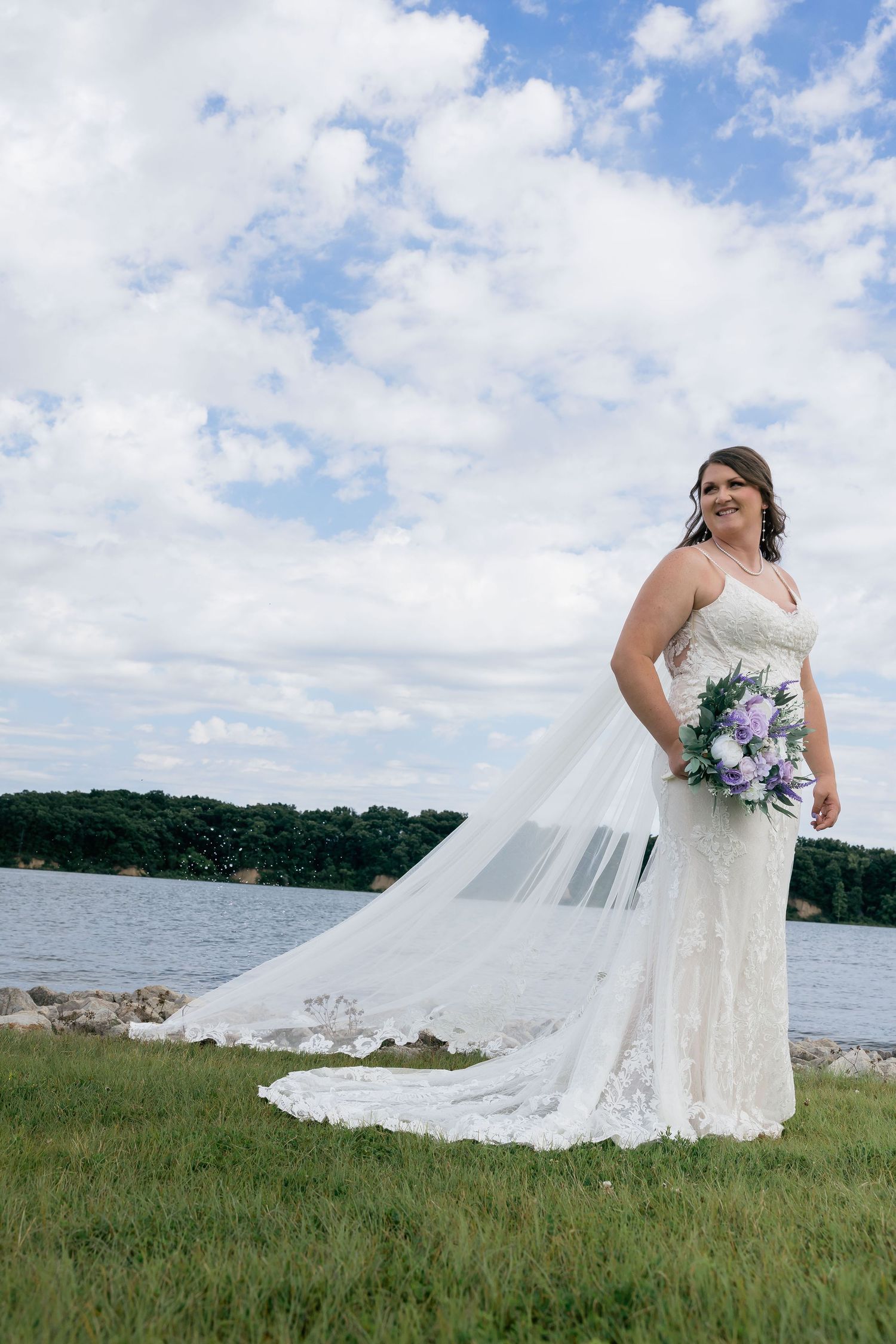 Bride in lace mermaid gown with flowing veil holds purple bouquet, posing by lakeside under partly cloudy sky.