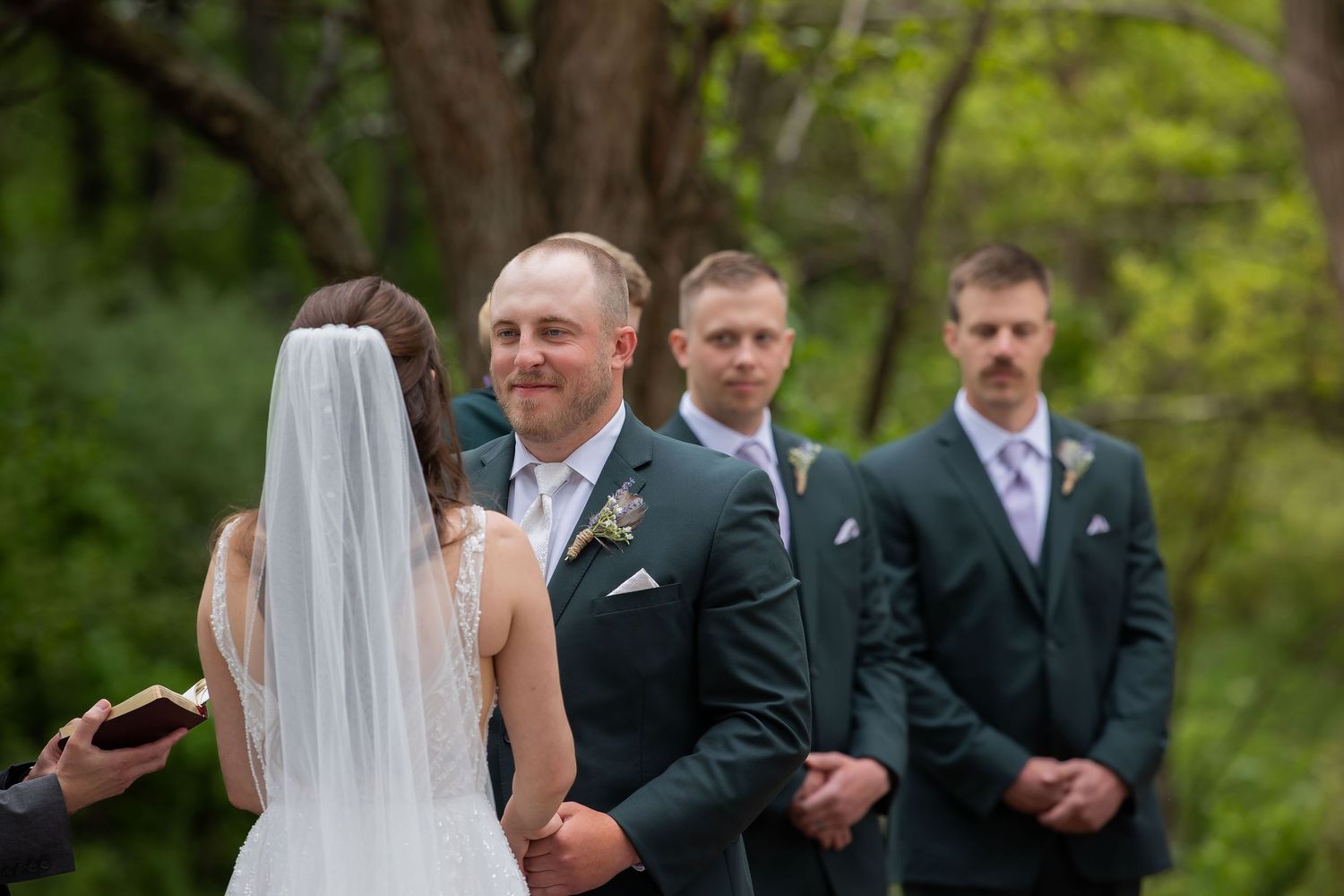 Groom smiles at bride during outdoor wedding ceremony with groomsmen in dark suits standing behind him.