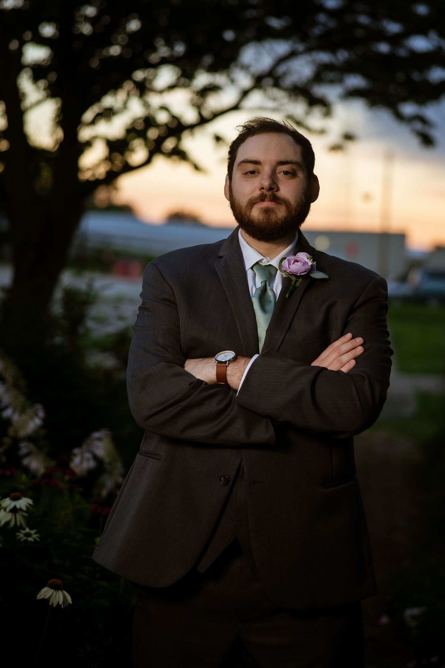A young man in a dark suit with boutonniere stands arms crossed at sunset during a formal outdoor event.