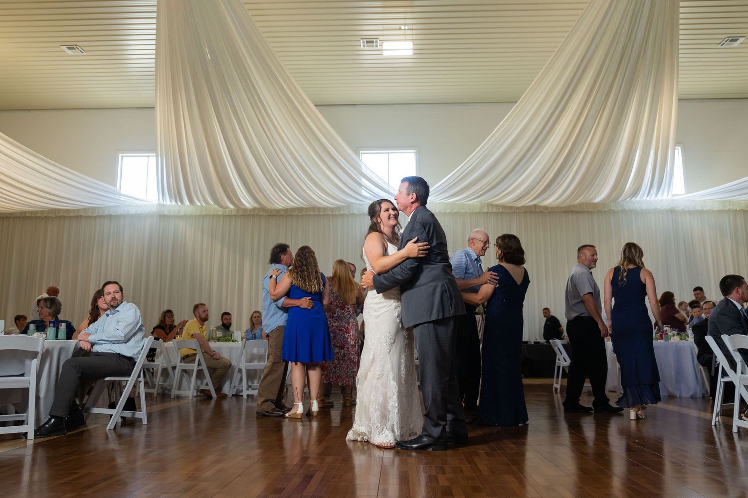Couple shares first dance at wedding reception in elegant white draped venue with guests watching.