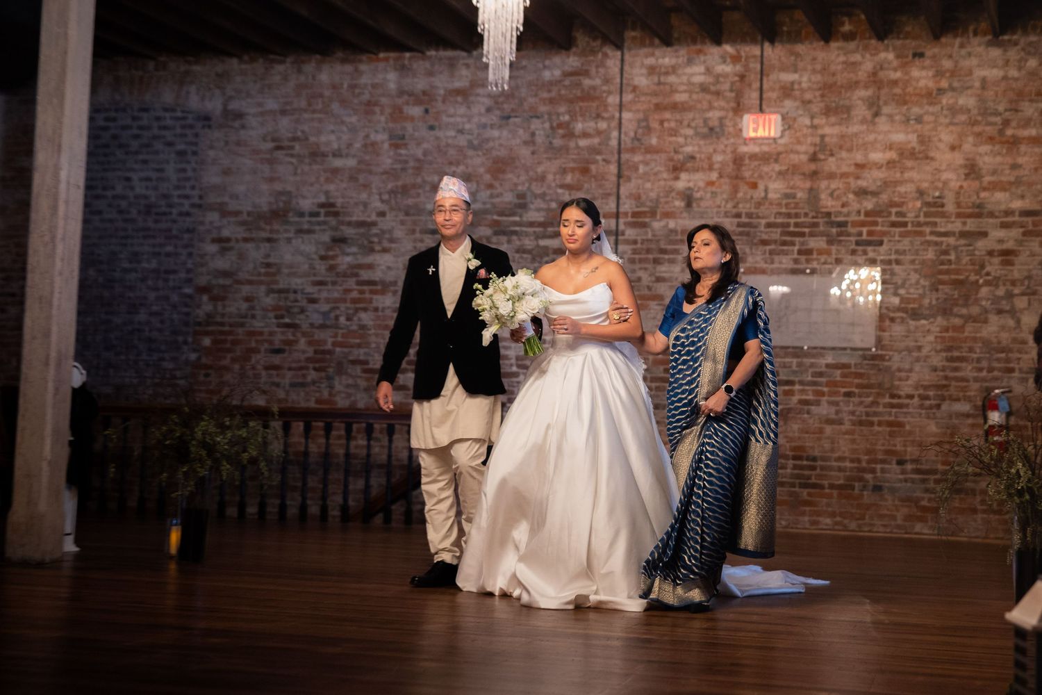Three people stand together at a wedding celebration in front of a rustic brick wall in an indoor venue.