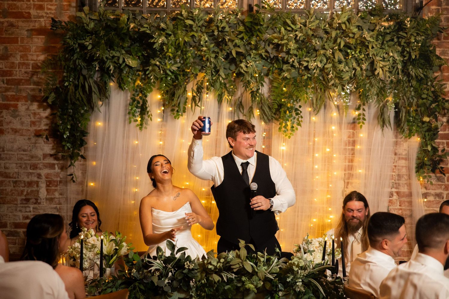 Wedding couple makes toasts at reception head table decorated with lush greenery and twinkle lights.