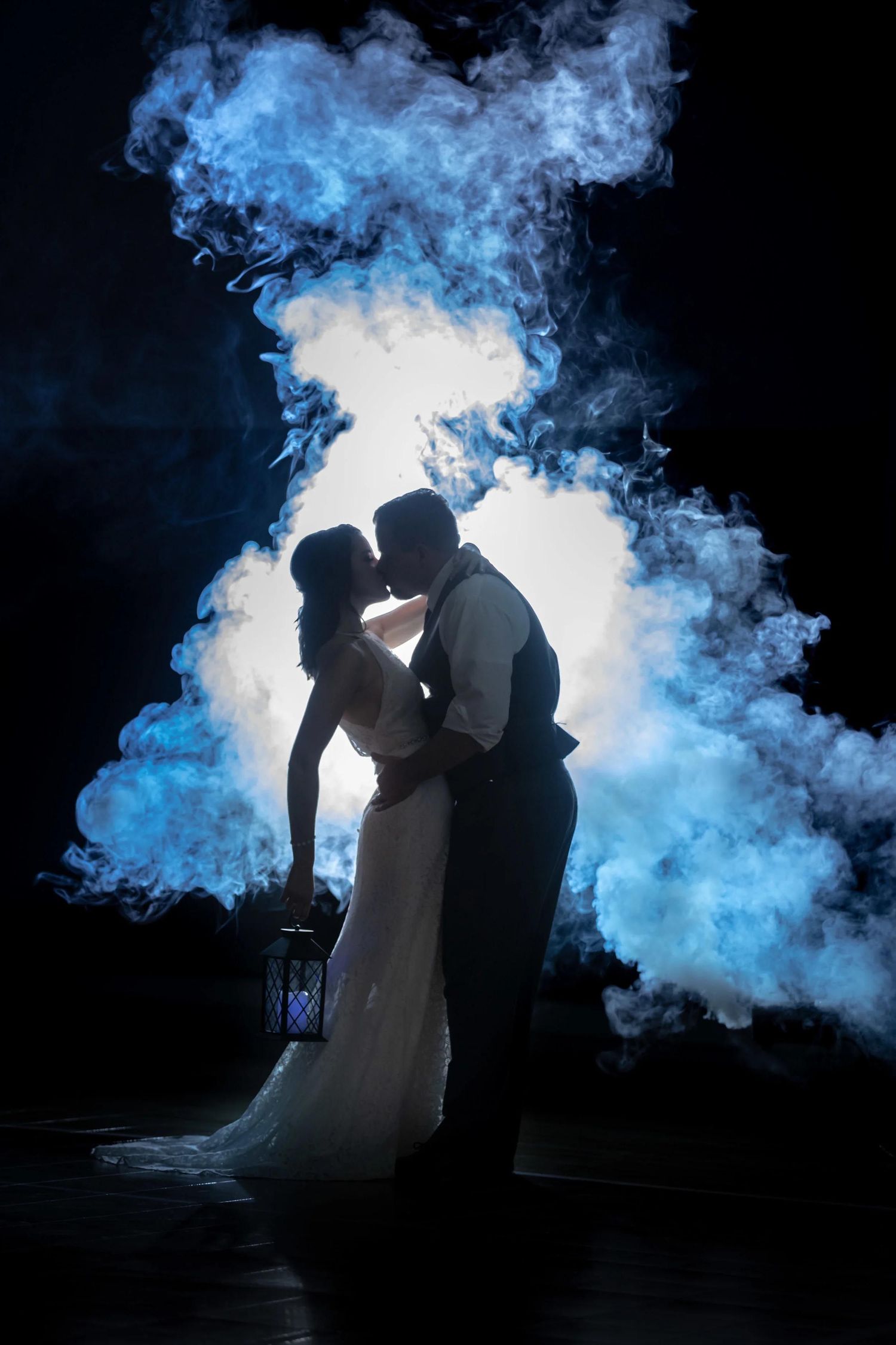 Bride and groom silhouette kissing surrounded by dramatic blue smoke clouds against dark background.