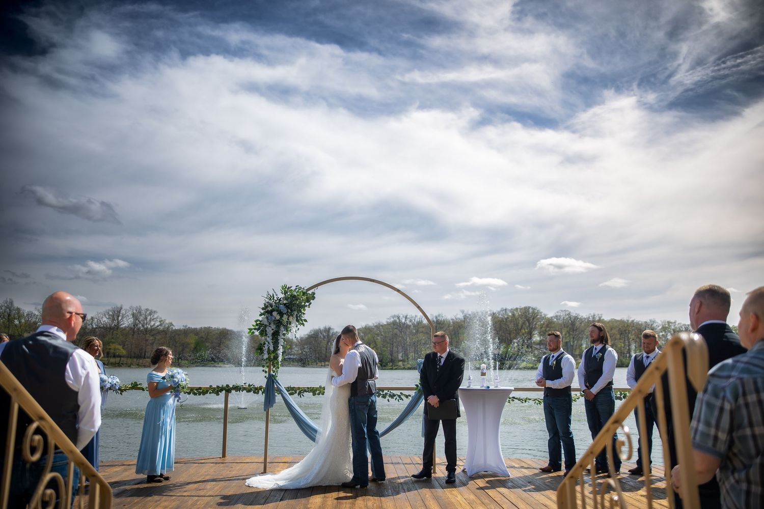 Bride and groom kiss at outdoor waterfront wedding ceremony under circular floral arch.