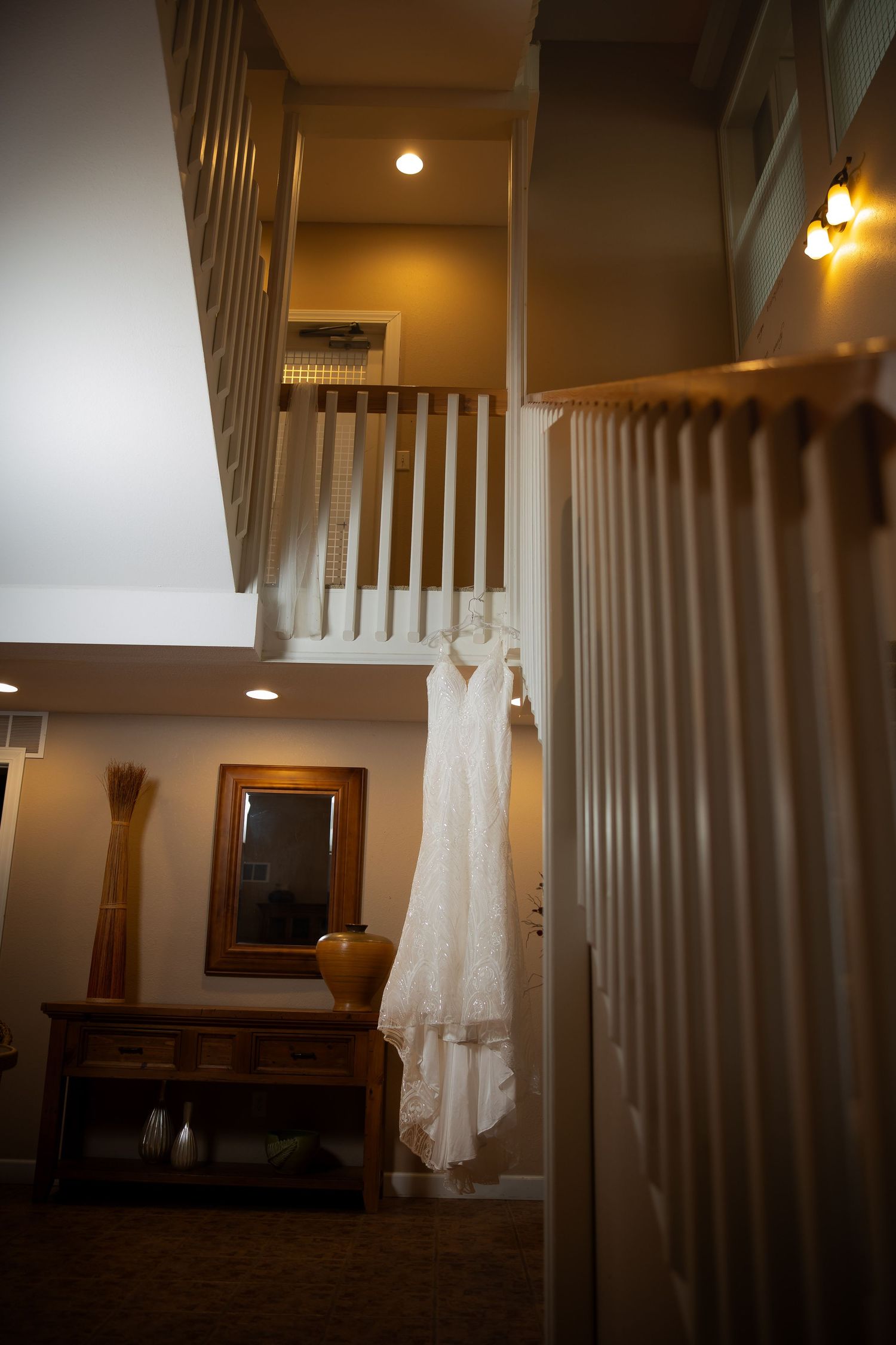 White lace wedding dress hanging from a balcony railing in an elegant warmly lit multi-level home interior.