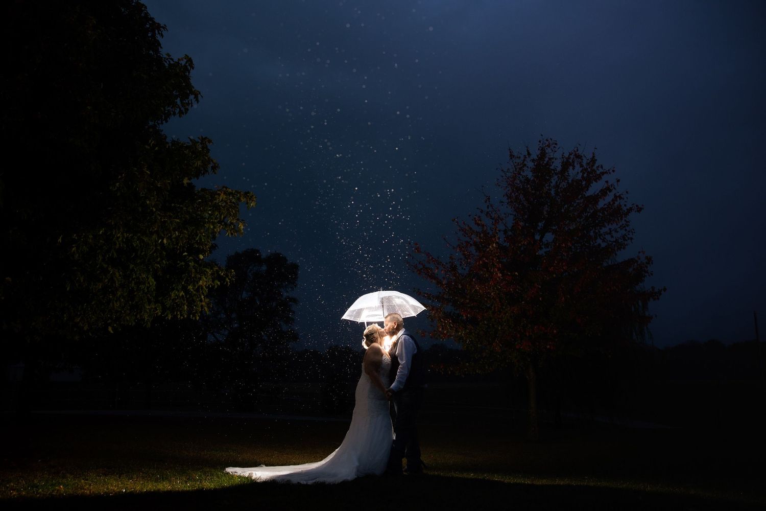 Bride and groom sharing a romantic kiss under a glowing umbrella in the rain at night.