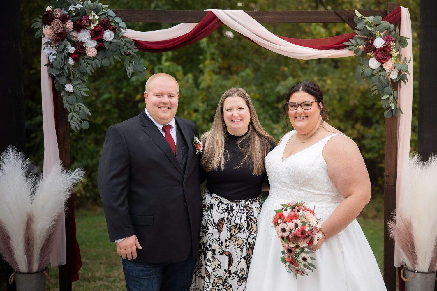 Wedding ceremony under a burgundy and pink floral arch with pampas grass decorations in an outdoor garden setting.