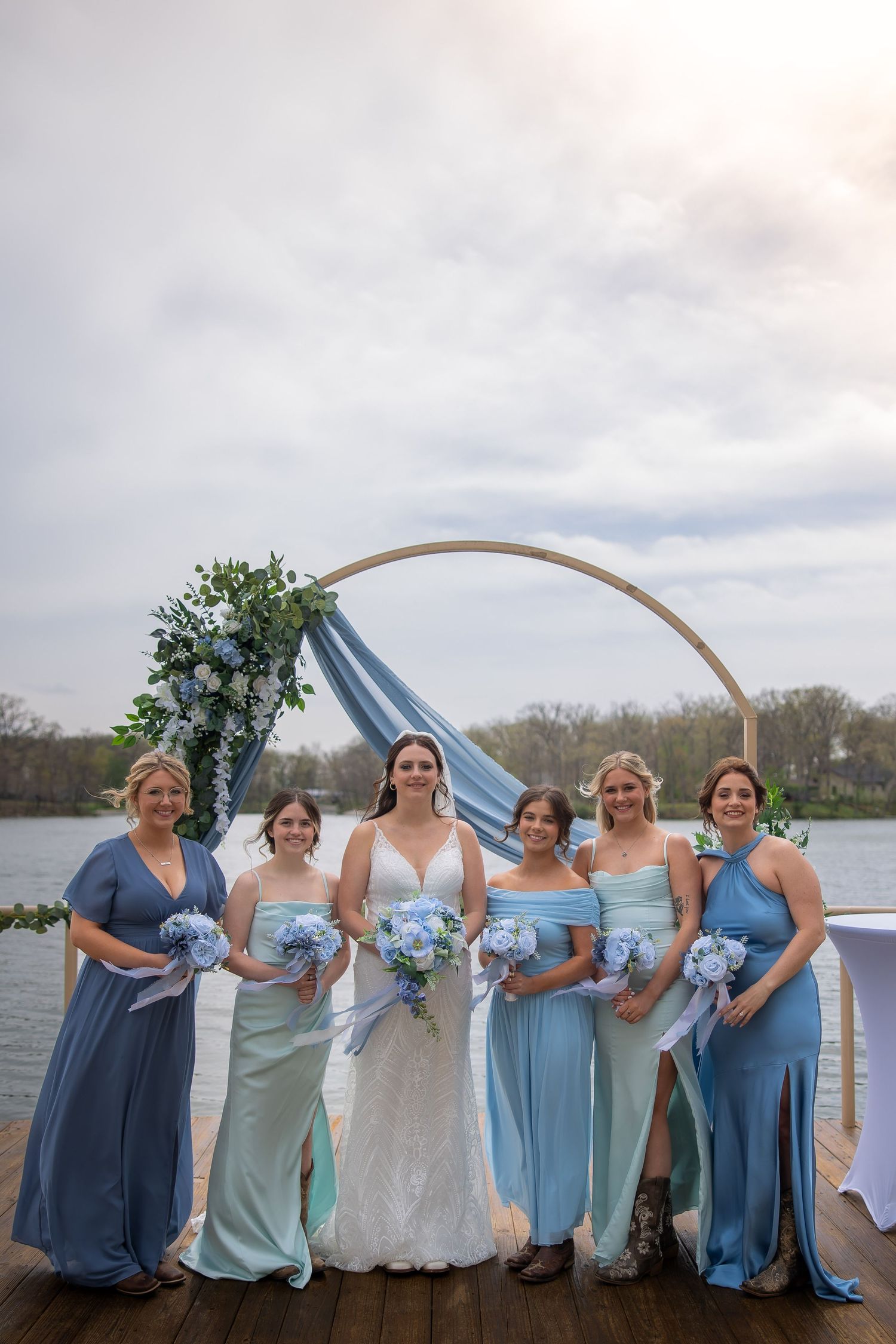 Bride and five bridesmaids in blue dresses holding bouquets pose by circular arch on waterfront dock.