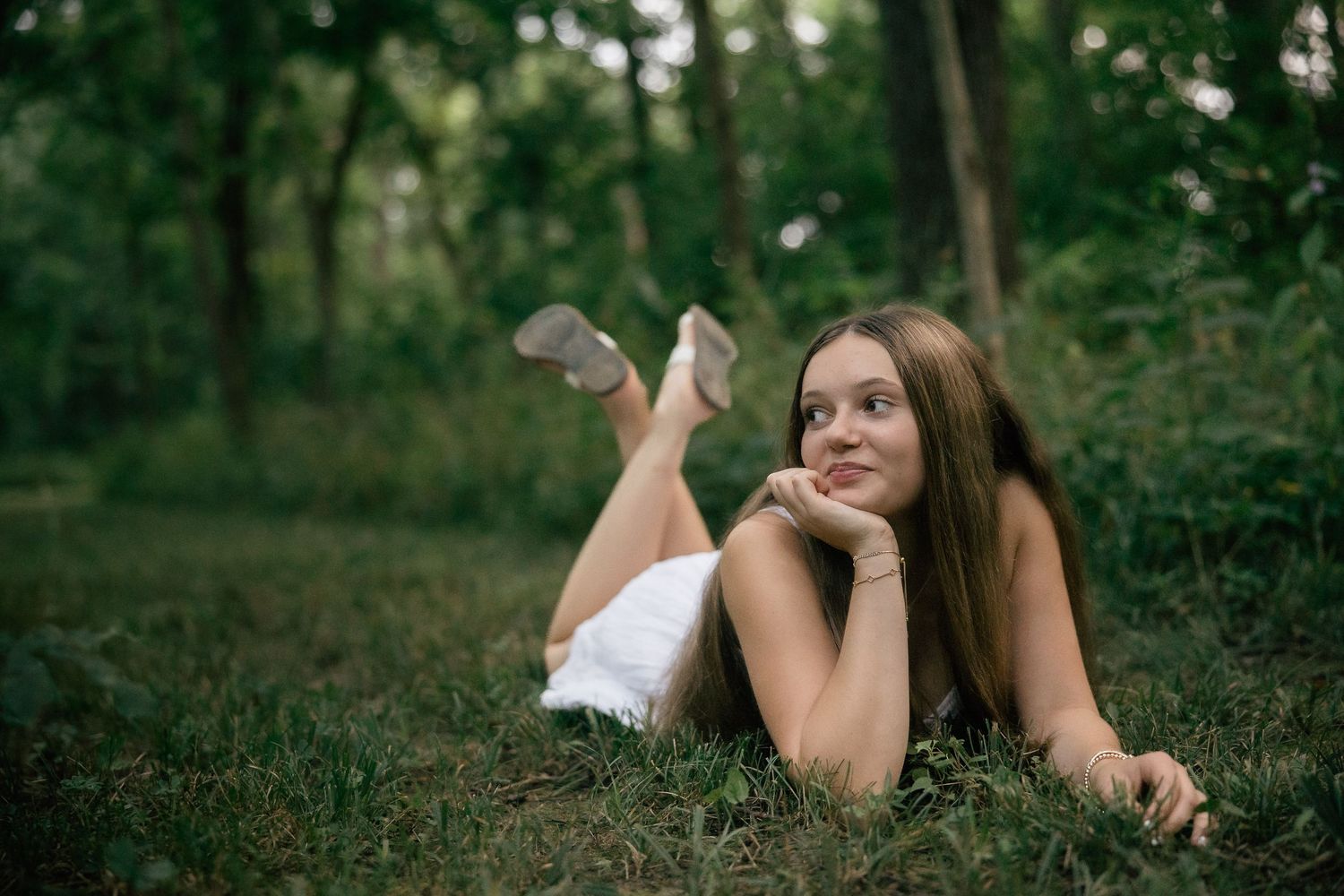 A person in white clothing lies relaxed on grass in a wooded setting.
