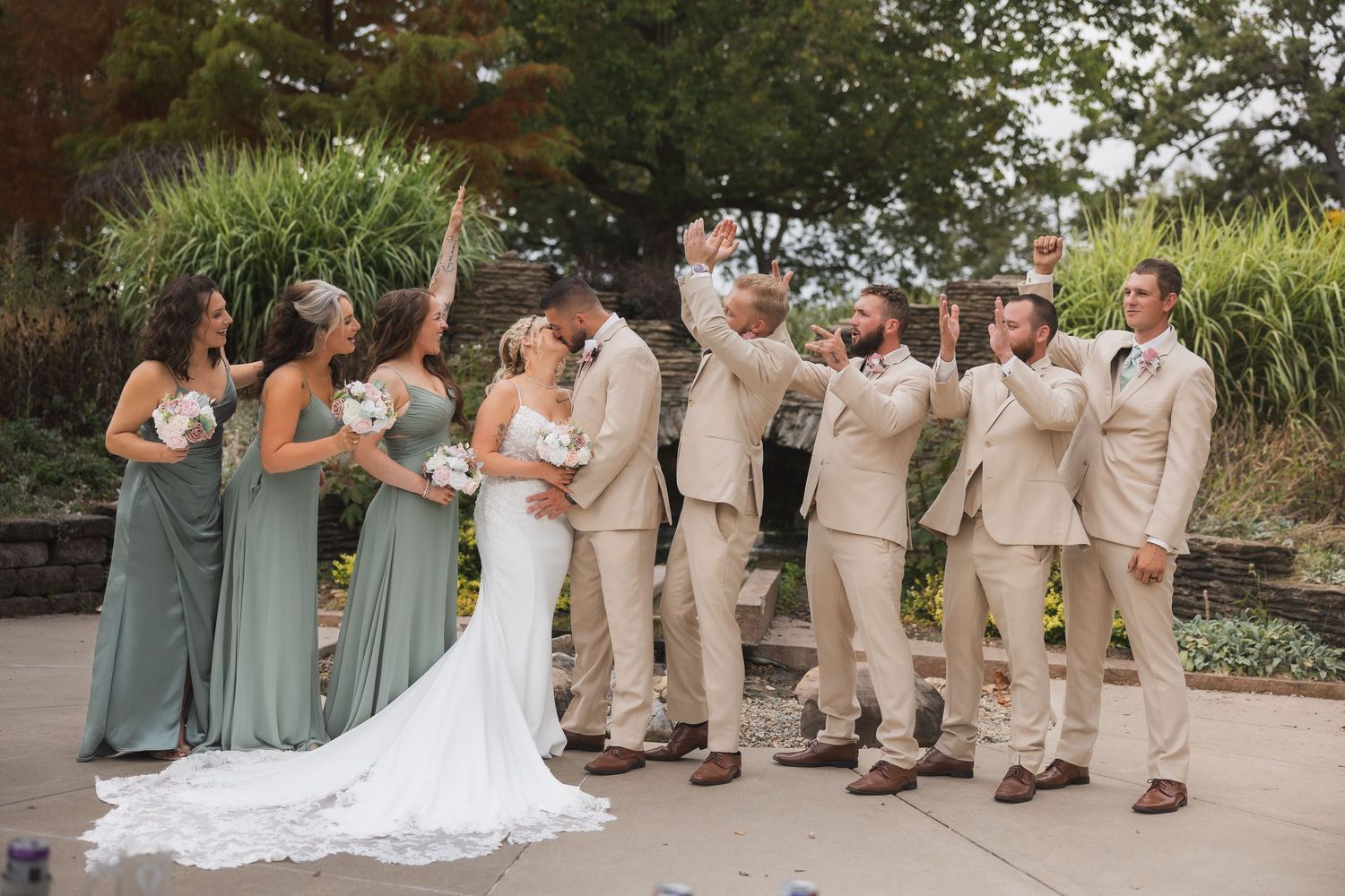 Wedding party in sage green dresses and tan suits posing joyfully outdoors with the bride in a white gown.