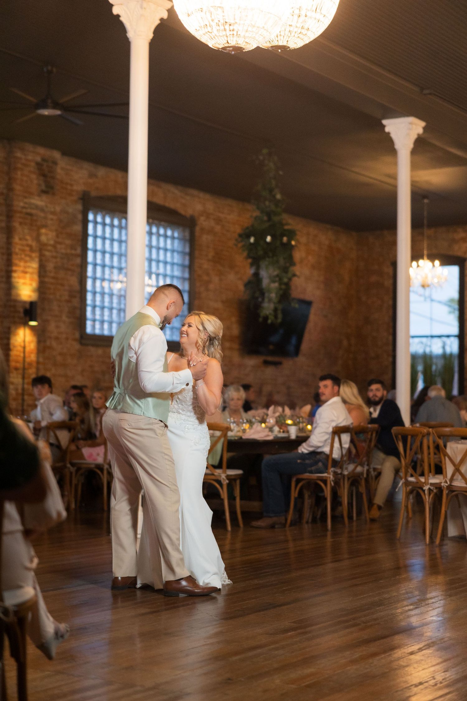 Couple shares first dance on hardwood floor under crystal chandelier in rustic brick reception venue.