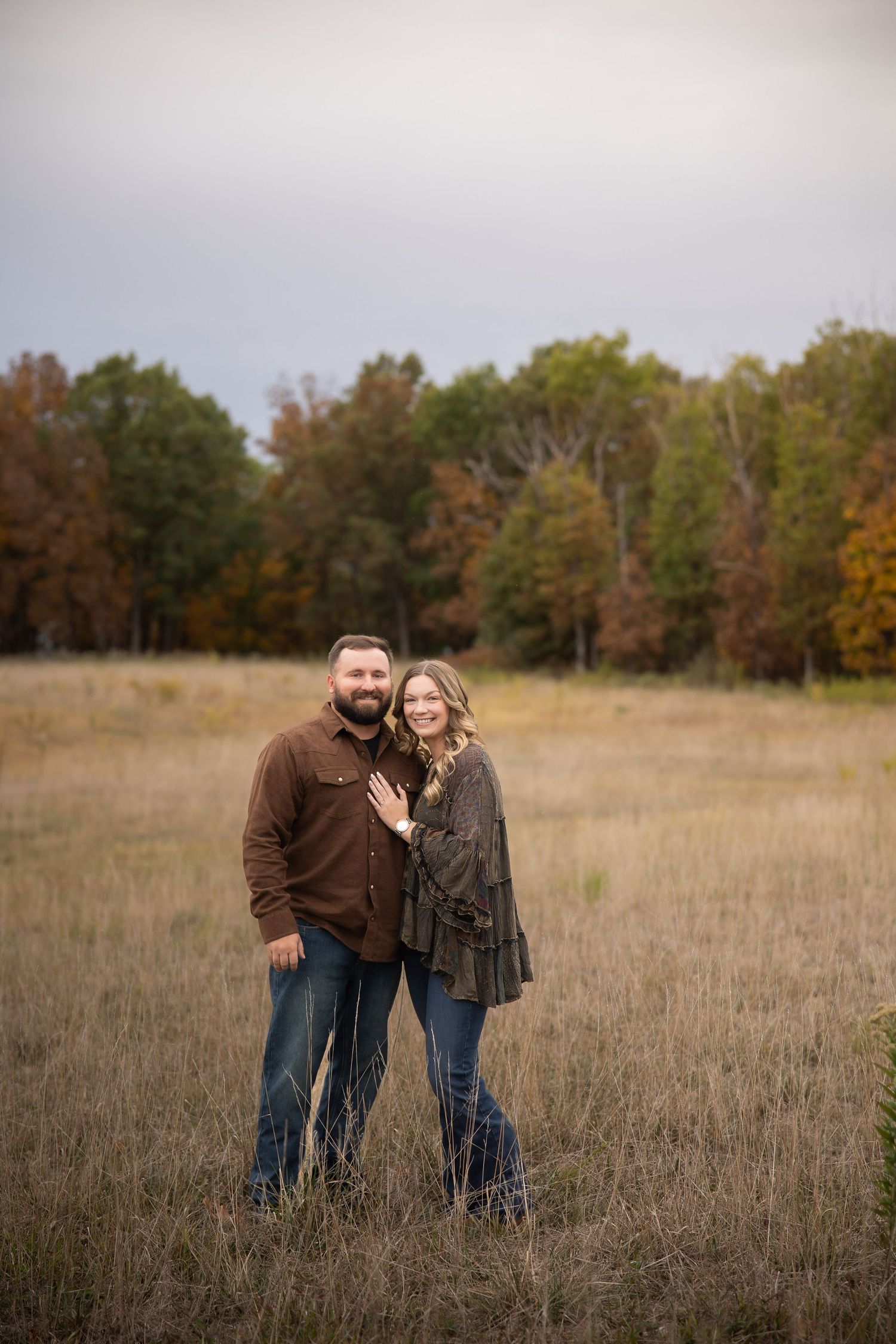 A couple stands together in a golden autumn field with fall foliage and trees in the background.