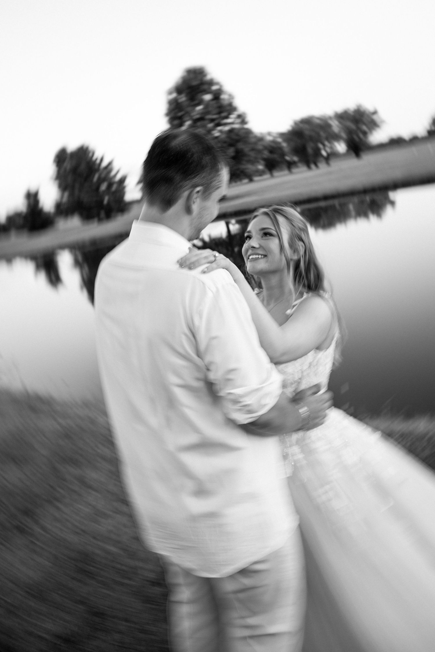 Black and white photo of smiling bride and groom embracing near reflective lake at sunset.